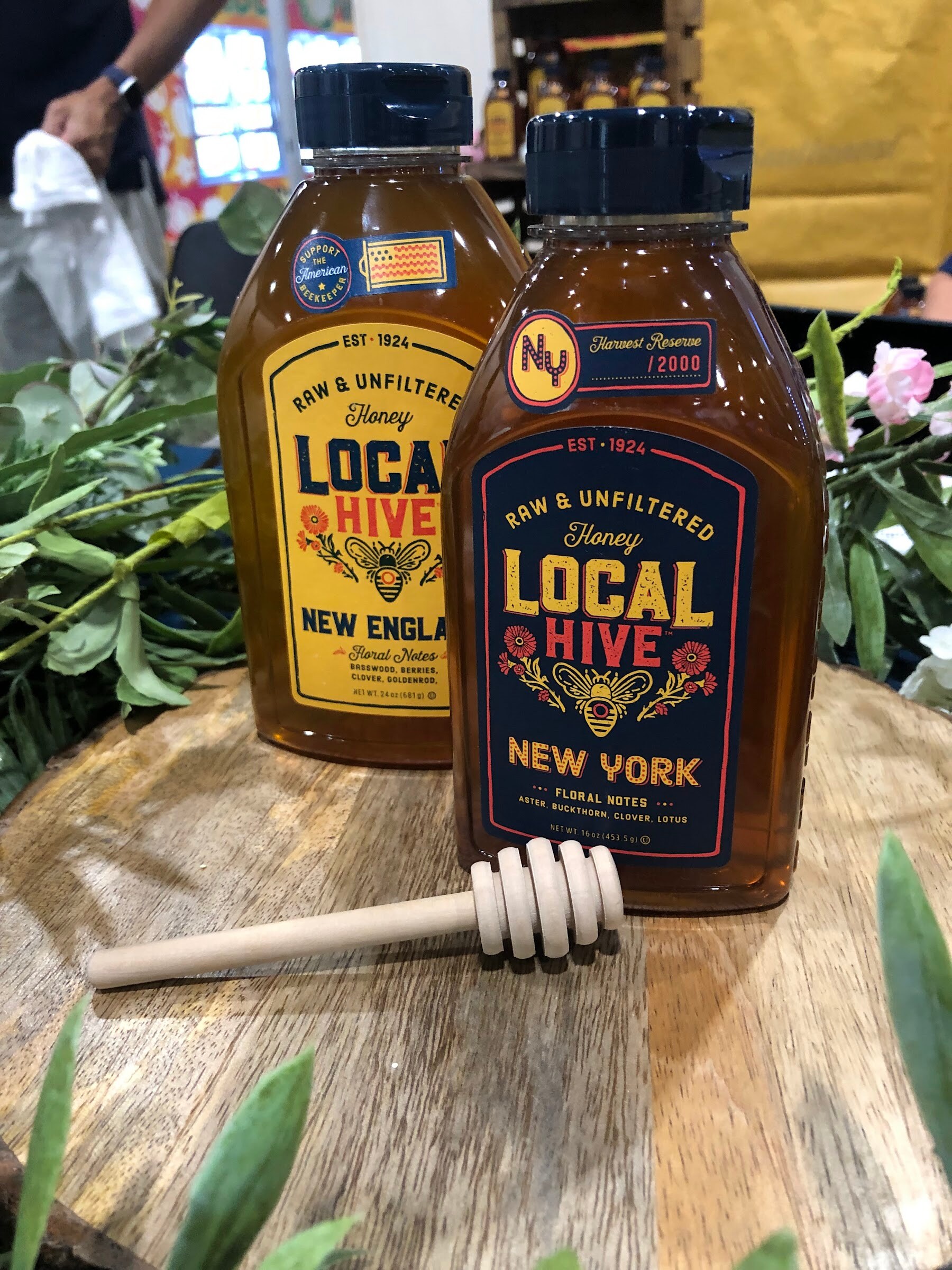 Two Local Hive raw and unfiltered honey squeeze bottles, New England and New York, on a wooden slice with a honey dipper