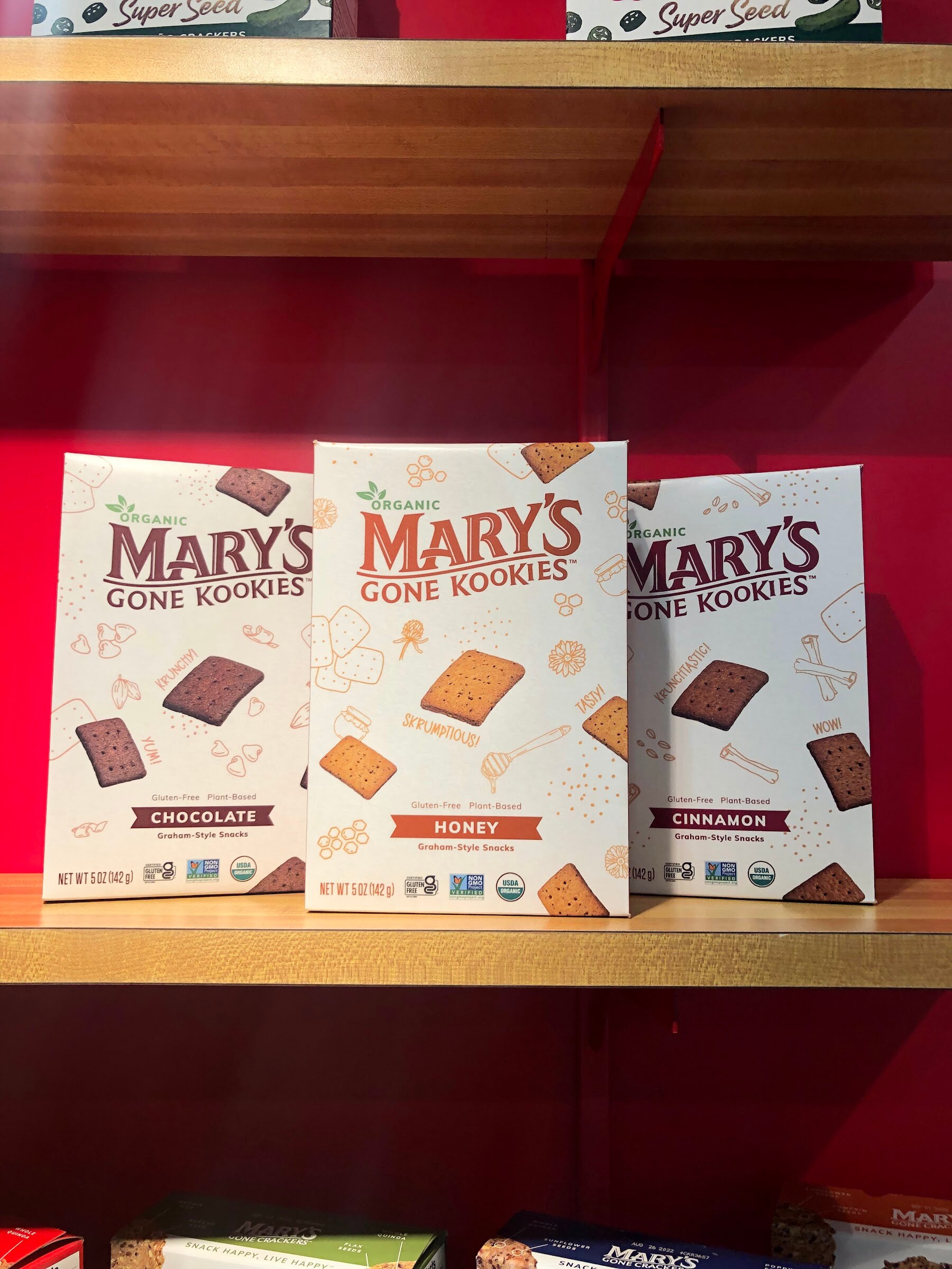 Three boxes of Mary's Gone Kookies in chocolate, honey and cinnamon flavors on a wooden shelf against a red wall
