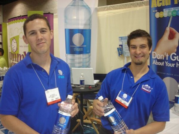 Two smiling men in blue polo shirts and badge lanyards each hold a bottle of Evamor water in front of a large Evamor water bottle banner.