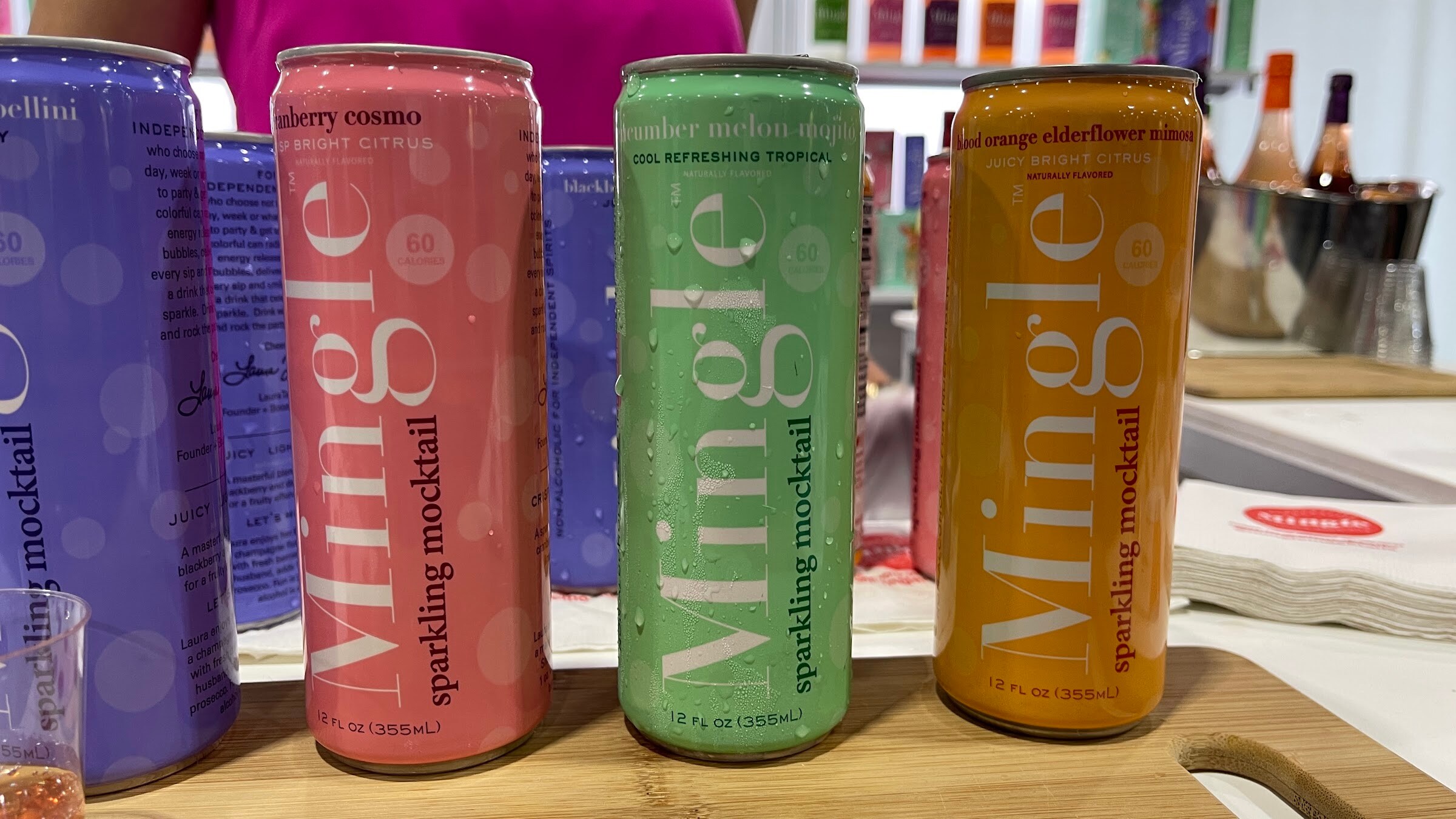 Three Mingle sparkling mocktail cans in pink, green and orange on a wooden board