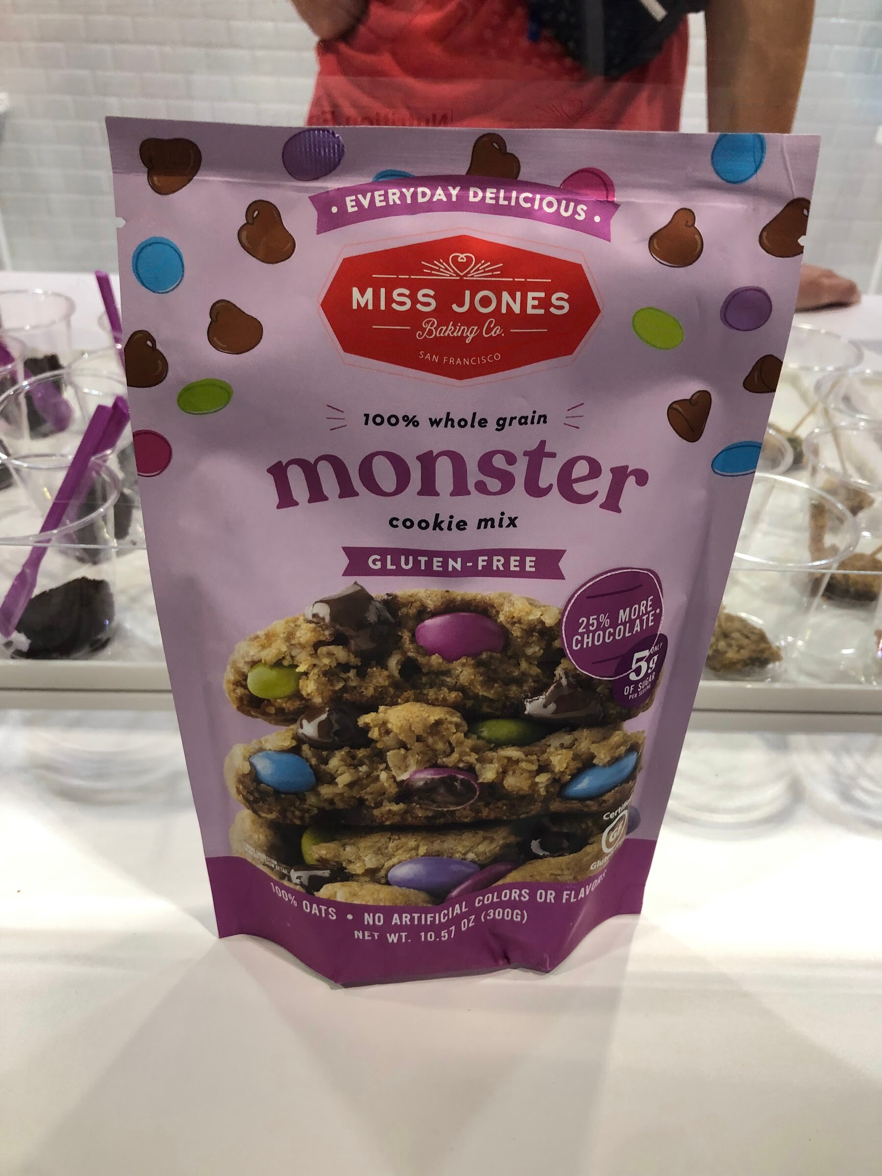 A purple Miss Jones Baking Co. monster cookie mix pouch on a counter with sample cups behind it