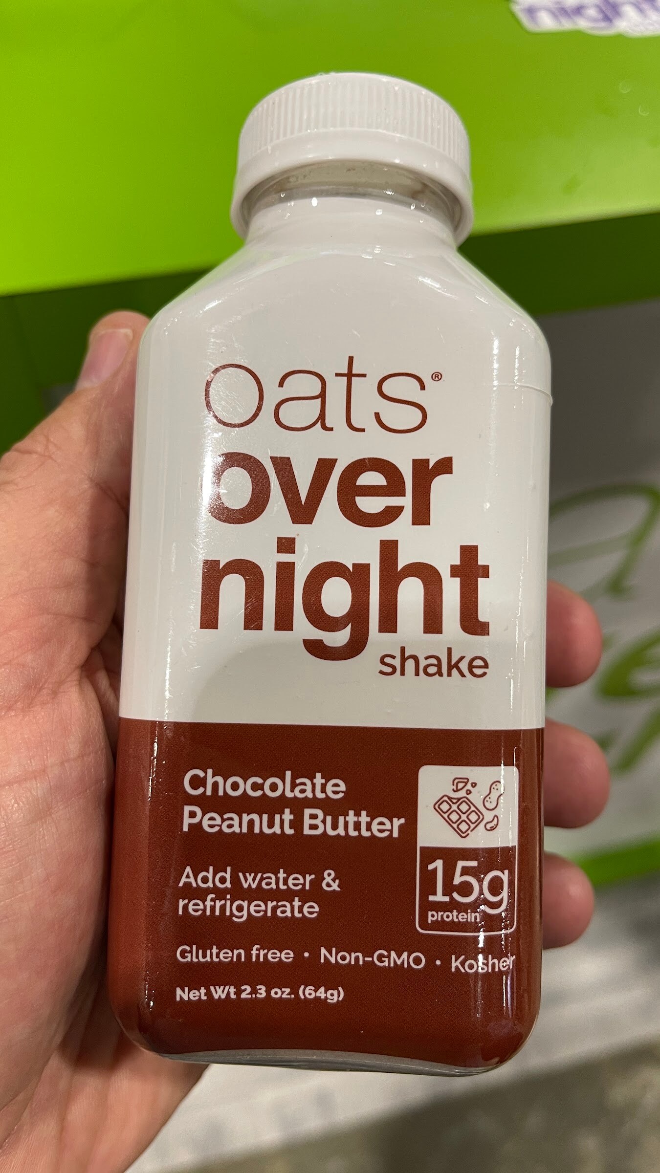 A hand holds an oats overnight chocolate peanut butter shake bottle in front of a green display