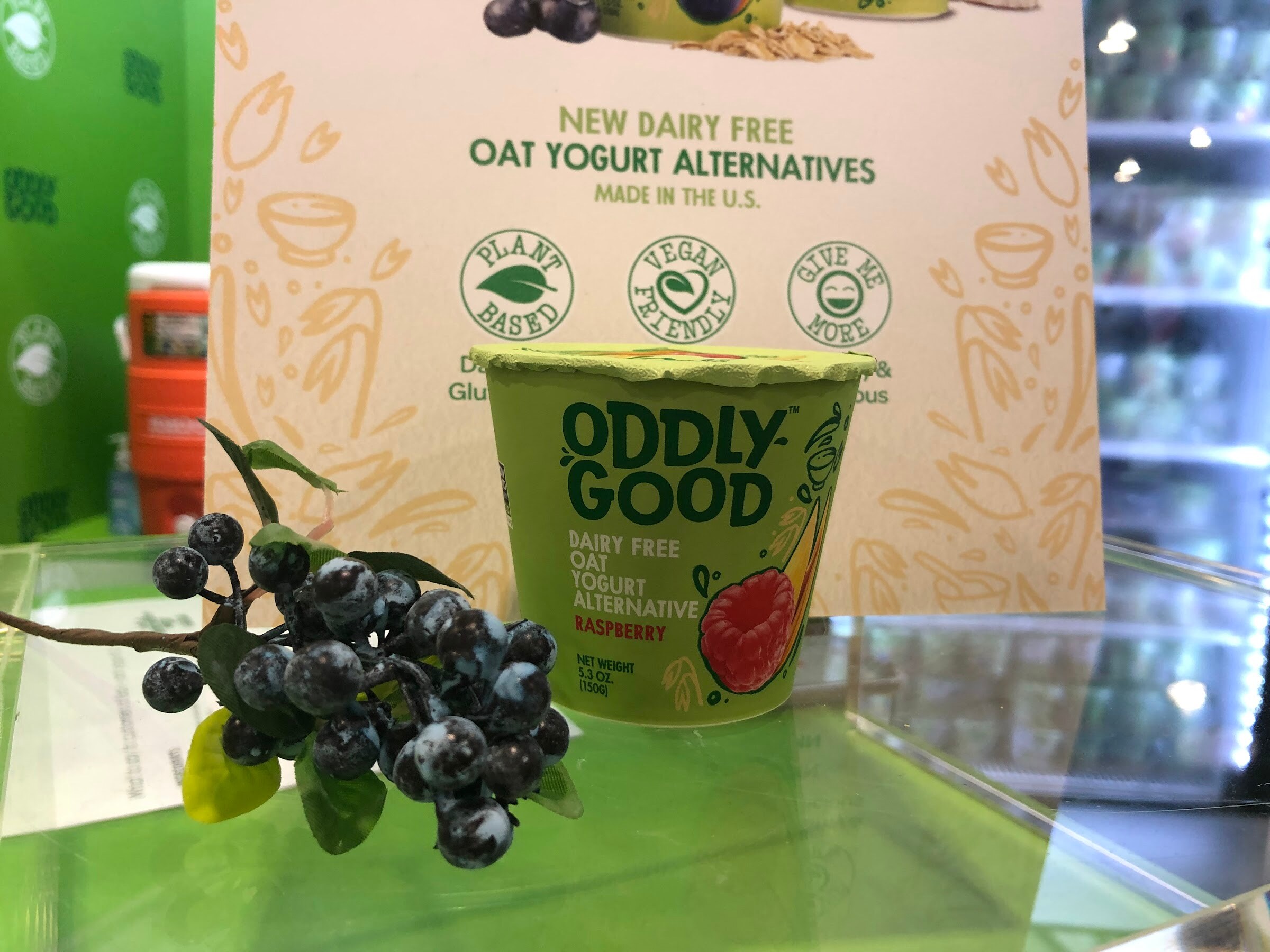 An Oddly Good raspberry oat yogurt cup on a green glass shelf in front of a sign for dairy-free oat yogurt alternatives