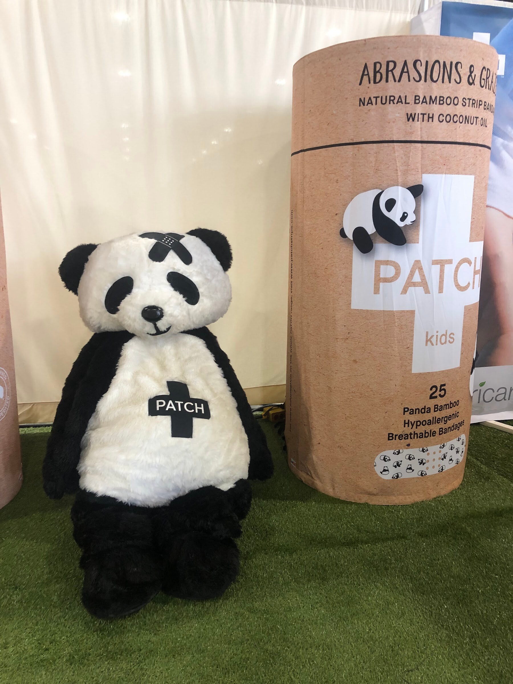 A stuffed panda wearing a Patch cross logo sits beside a tall tube of Patch kids bandages on artificial grass