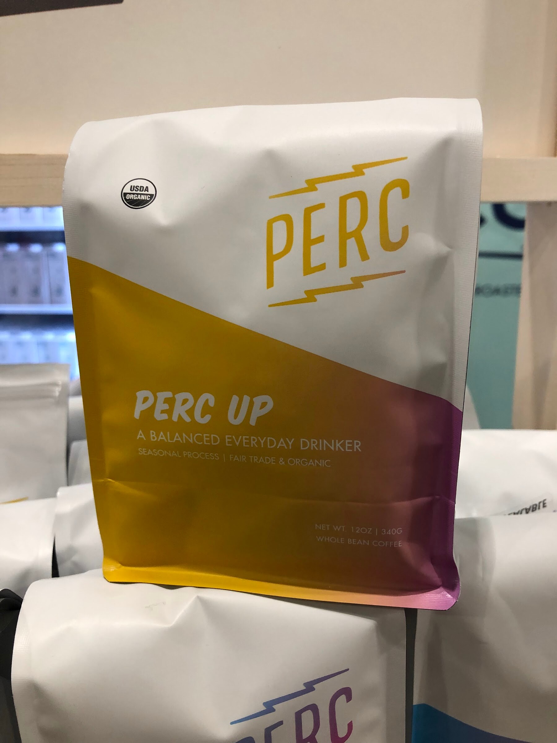A white, gold and purple Perc Up coffee bag stacked on other Perc coffee bags