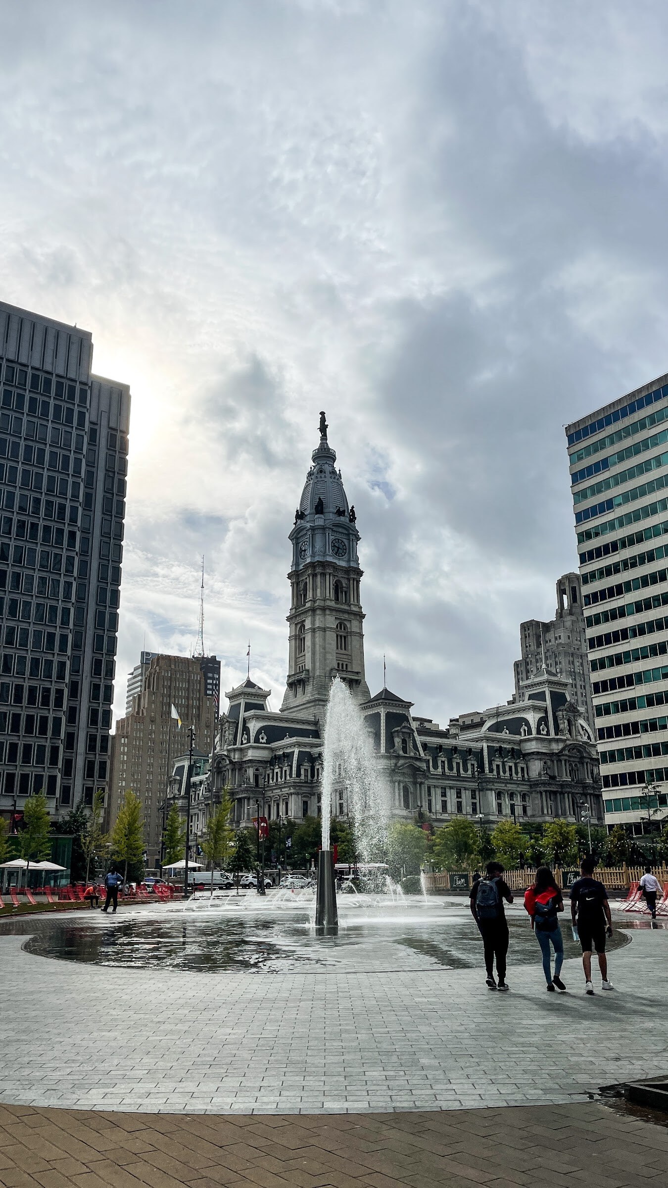 The Philadelphia City Hall tower rises over a plaza with a fountain and a few people under a cloudy sky