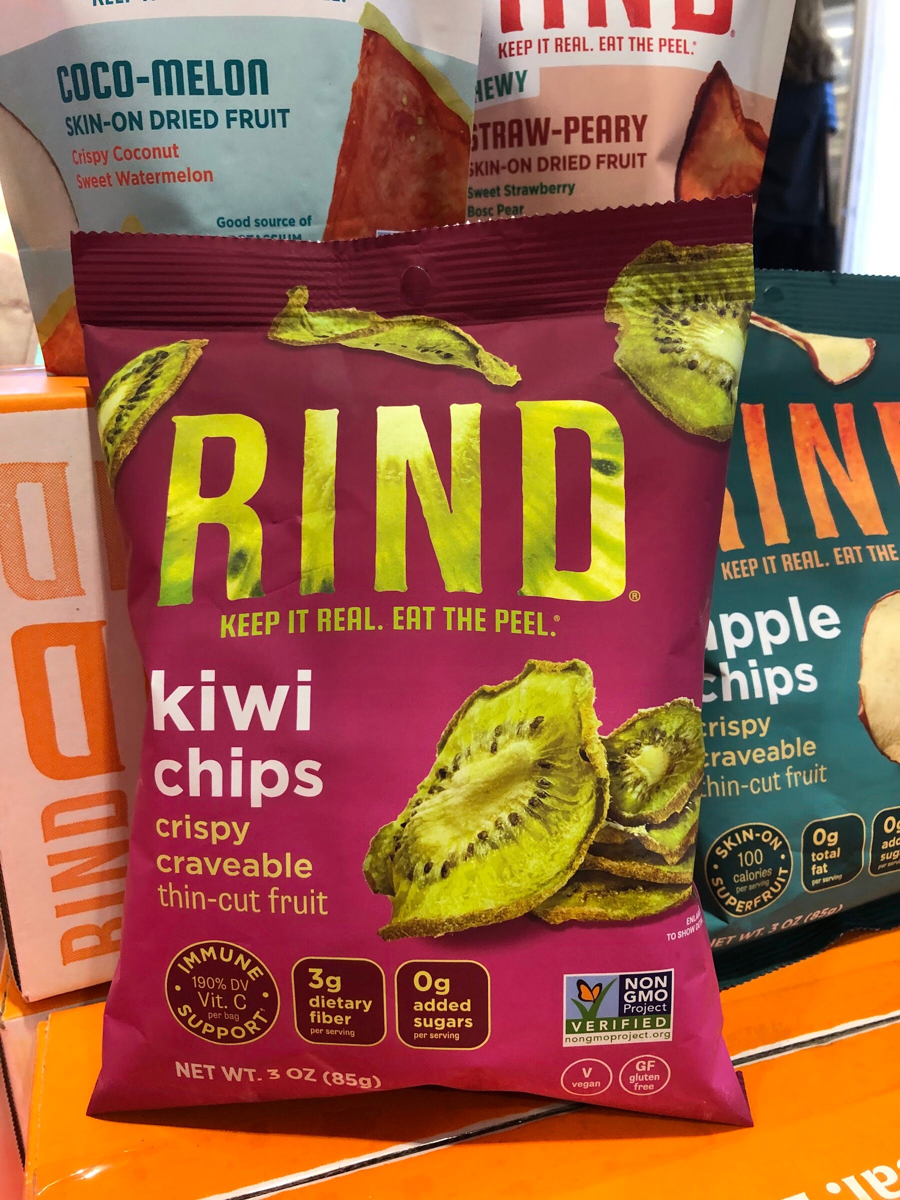 A magenta RIND kiwi chips bag in front of a bag of apple chips on an orange display