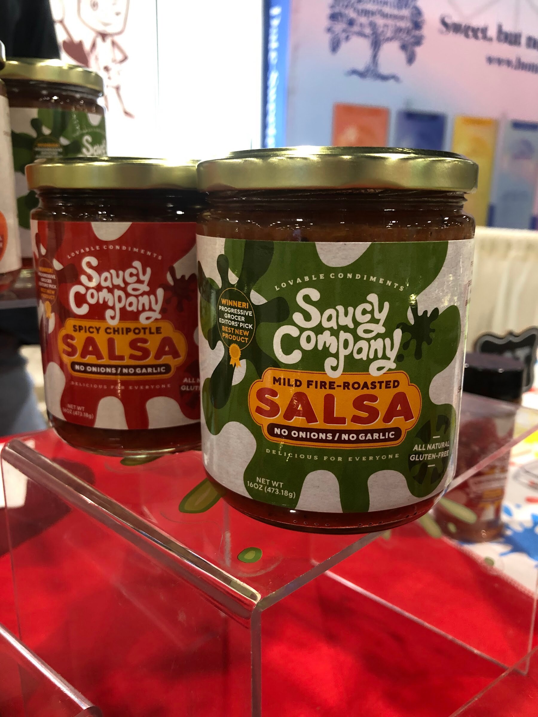 Jars of Saucy Company spicy chipotle and mild fire-roasted salsa on a clear stand over a red cloth