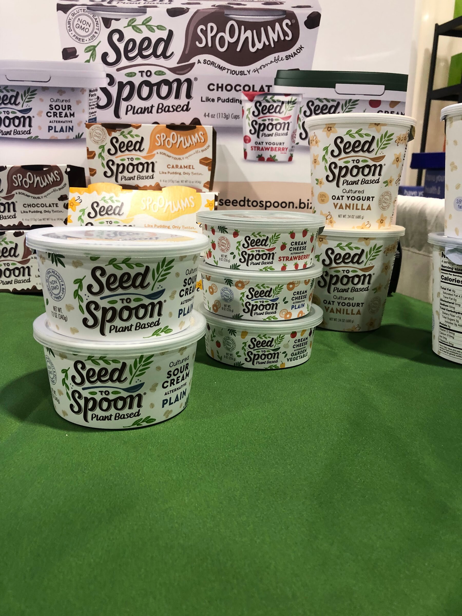 Seed to Spoon plant-based sour cream and yogurt cups stacked on green turf in front of a Spoonums display