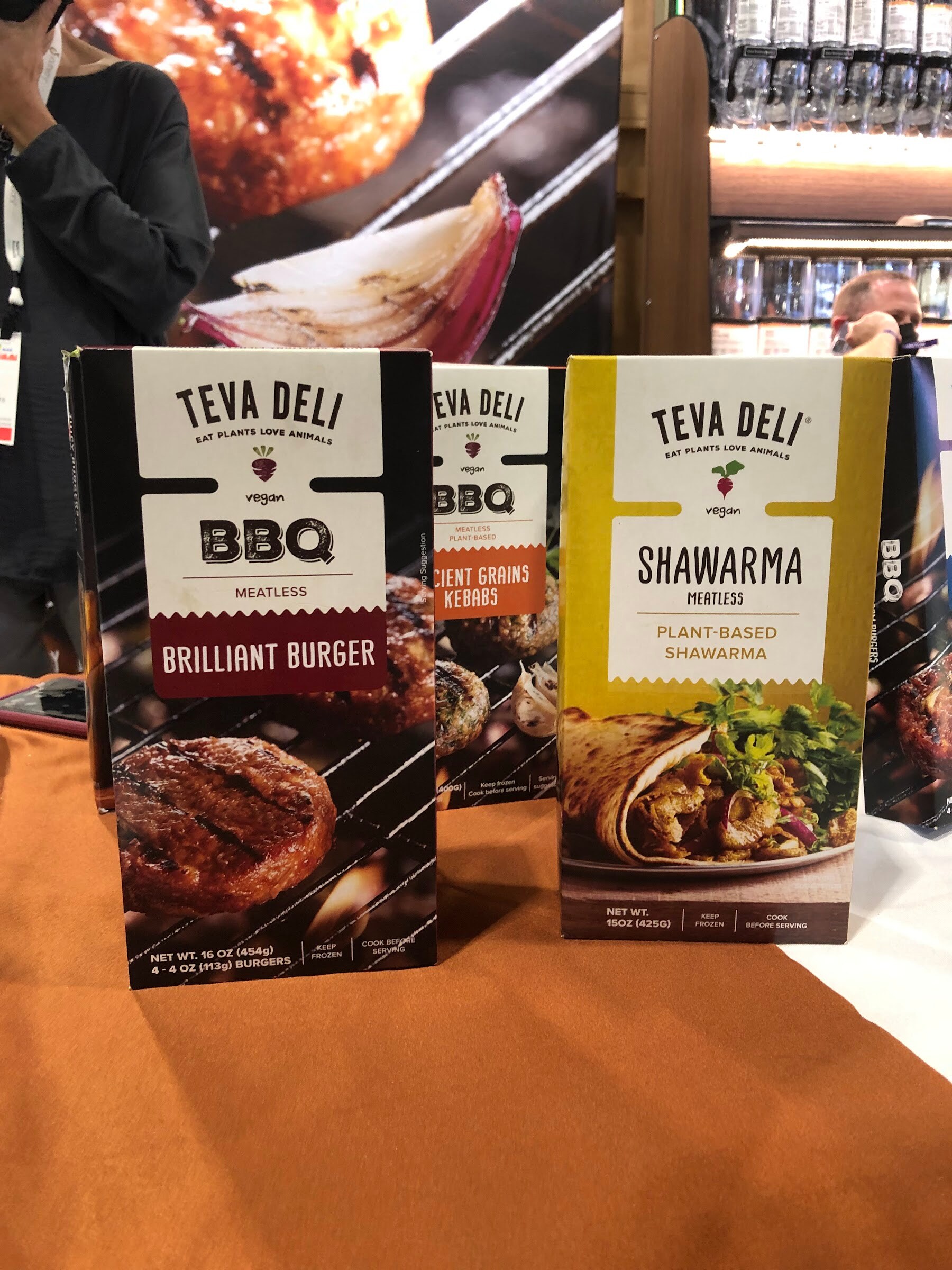 Teva Deli BBQ burger and shawarma boxes on an orange table with an exhibitor behind them