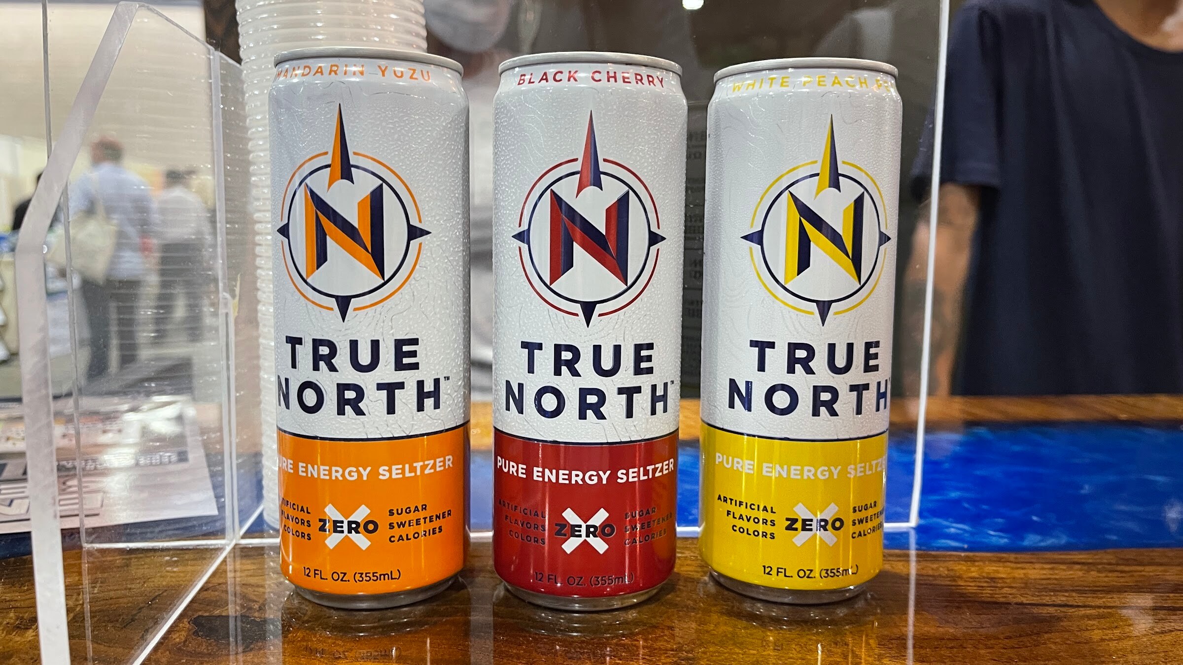 Three True North pure energy seltzer cans in orange, red and yellow behind a clear acrylic stand