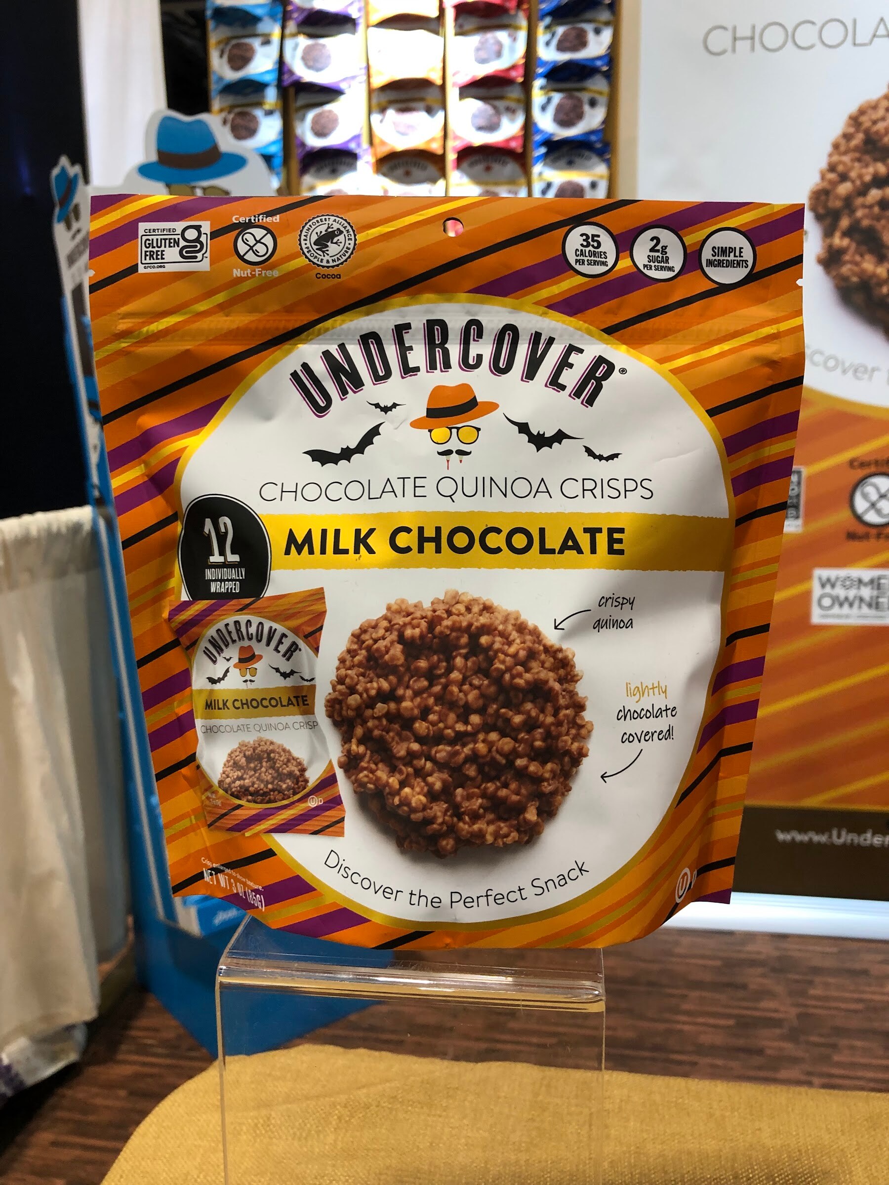 An Undercover milk chocolate quinoa crisps bag on a clear stand in front of a gold tablecloth