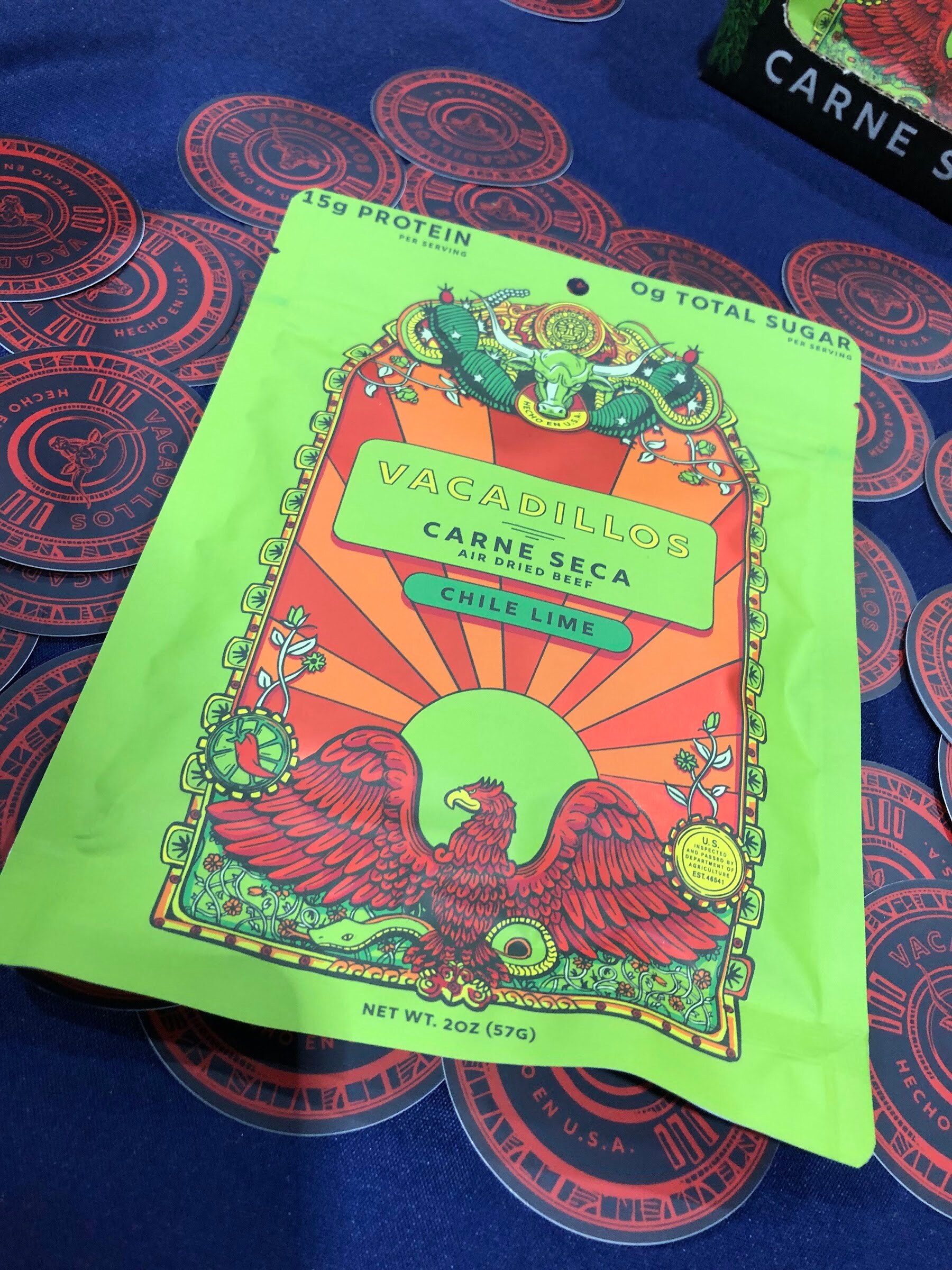 A bright green Vacadillos chile lime carne seca pouch with an eagle illustration on a blue tablecloth