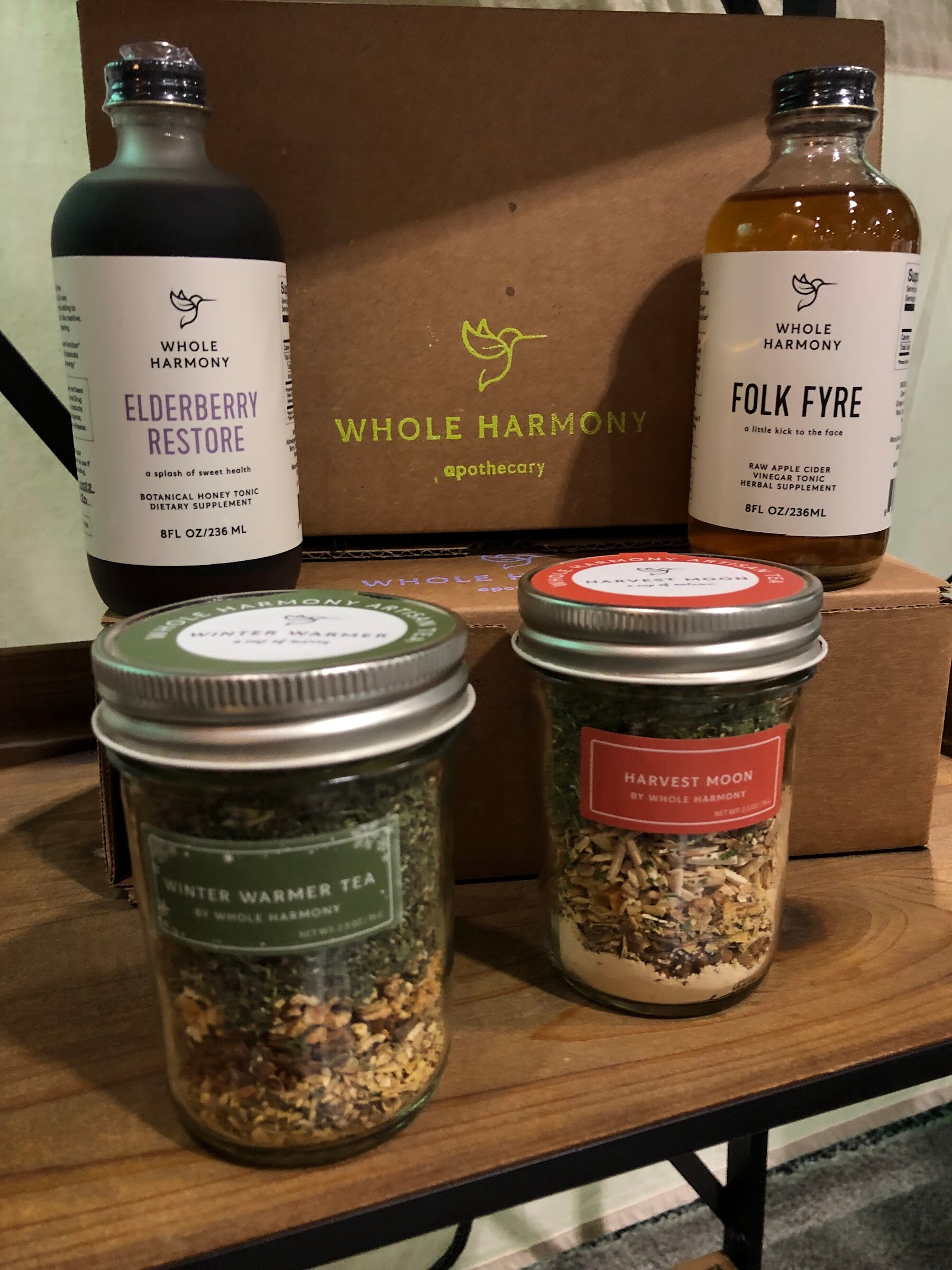 Whole Harmony Elderberry Restore and Folk Fyre bottles with Winter Warmer and Harvest Moon tea jars on a wooden stand