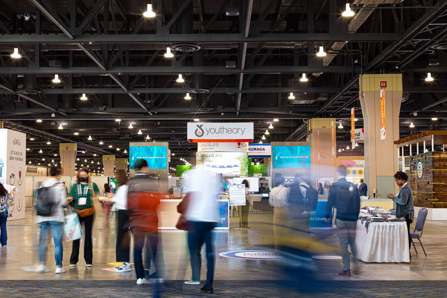 Motion-blurred attendees walk through a wide convention hall aisle lined with booths under high industrial ceiling lights
