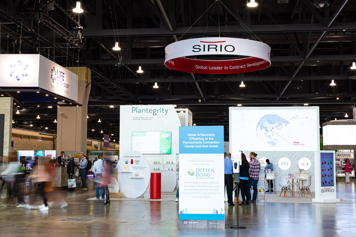 A wide view of the exhibit hall floor with Plantegrity and Sirio booths, a hanging ring sign and a few attendees standing between booths