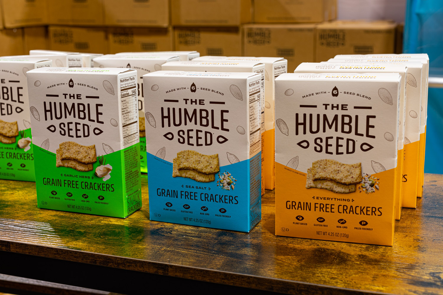 Three boxes of The Humble Seed grain free crackers in green, blue and orange stand on a wooden shelf