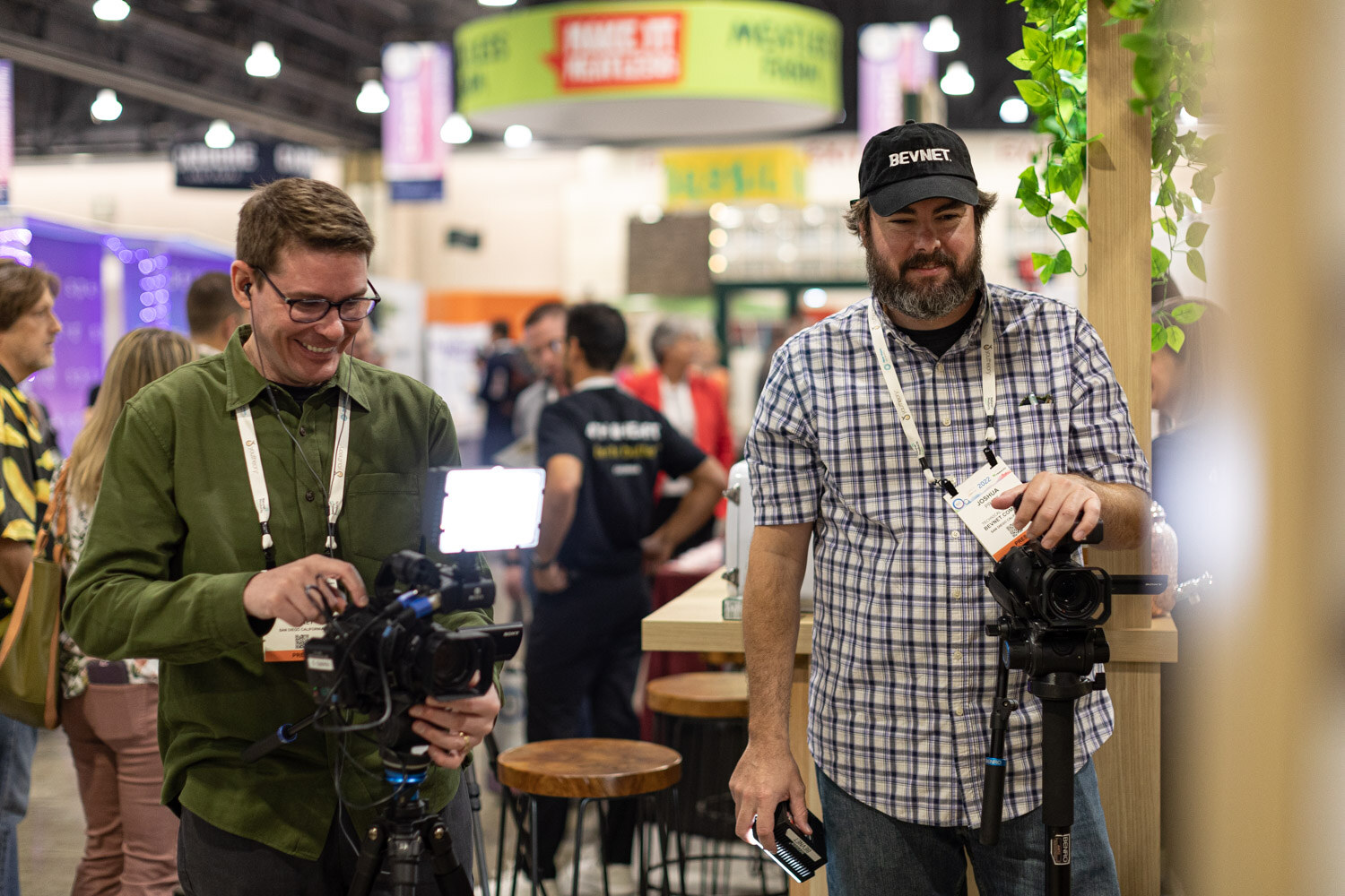 Two men with badges and a video camera on a tripod smile near a wooden booth with bar stools while the exhibit hall bustles behind them