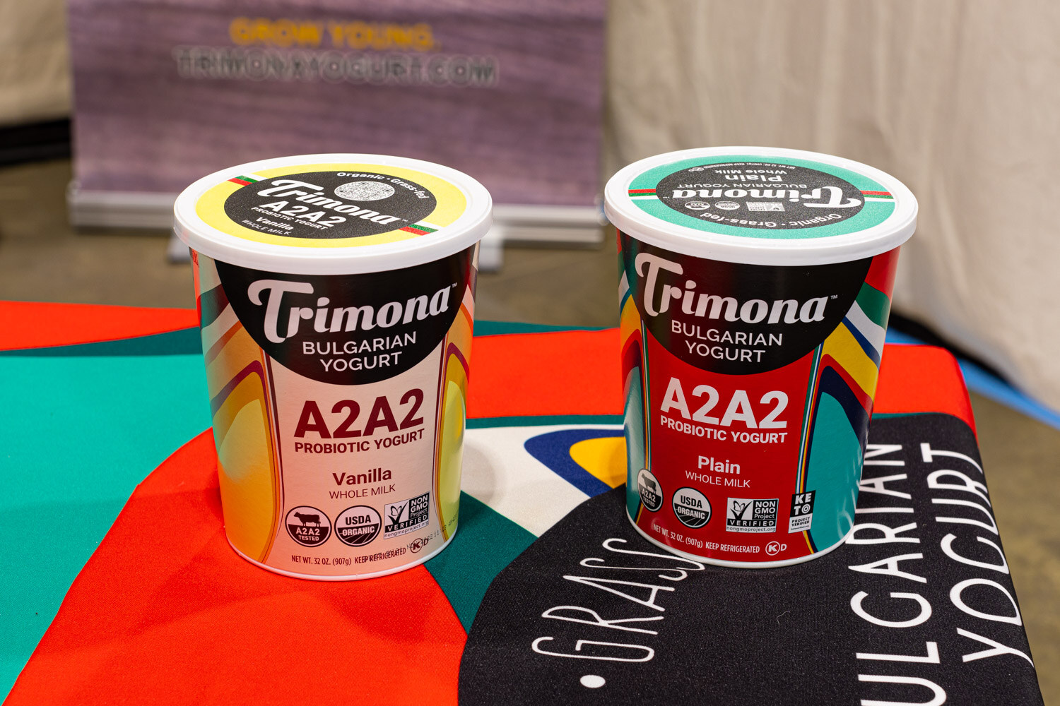 Two Trimona A2A2 probiotic Bulgarian yogurt cups, vanilla and plain, sit on a brightly colored table