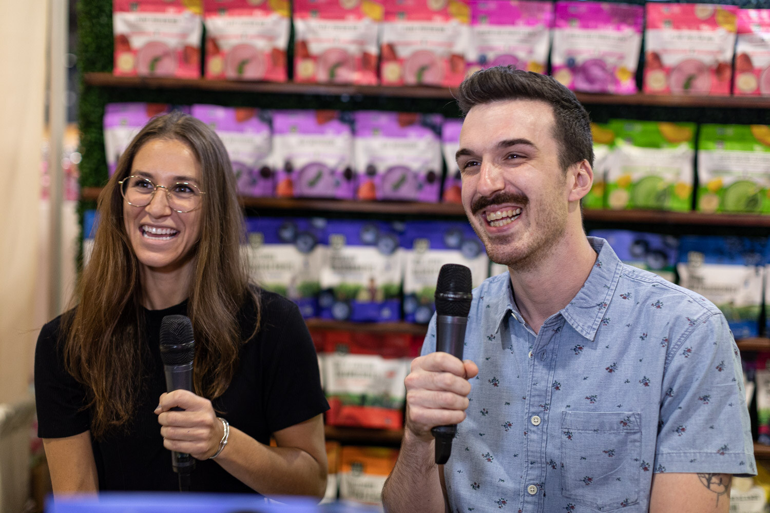 A woman and a man hold microphones and laugh during a video interview in front of shelves of purple, pink and green snack bags