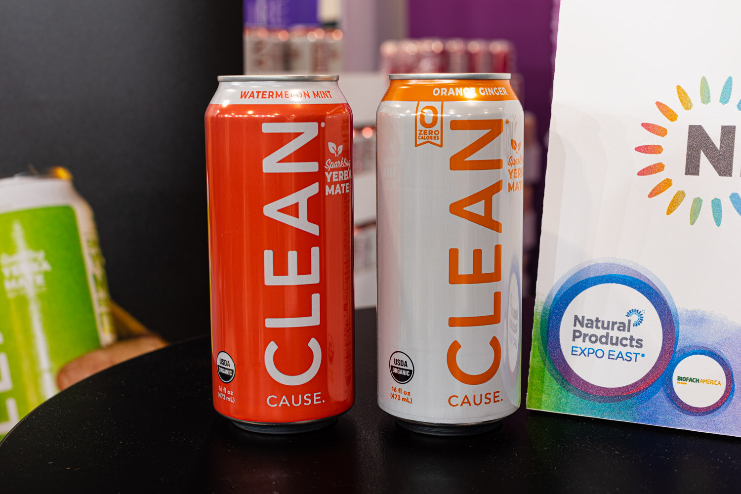 Red watermelon mint and white orange ginger CLEAN Cause cans stand next to a Natural Products Expo East sign