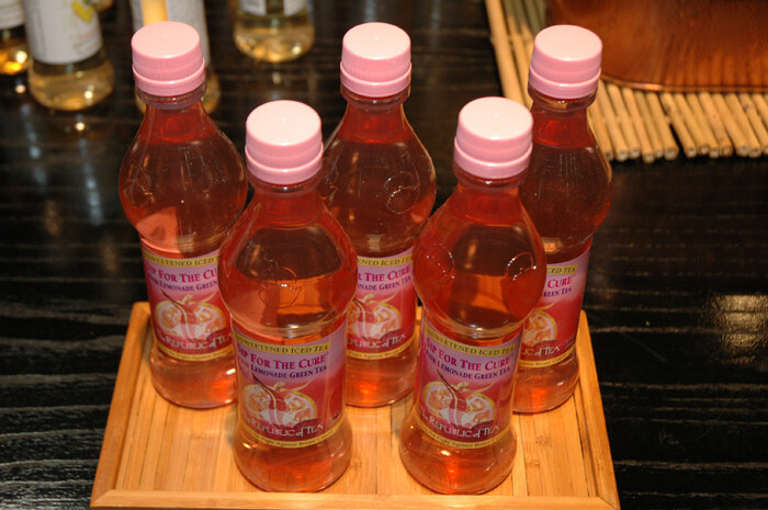 Five pink-capped bottles of The Republic of Tea sweetened iced tea with pink labels sit on a small bamboo tray on a dark table.