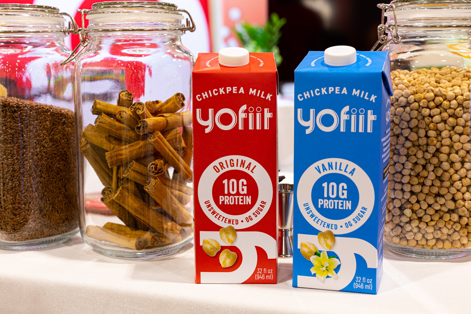 Red and blue Yofiit chickpea milk cartons in Original and Vanilla stand in front of glass jars of cinnamon sticks and beans