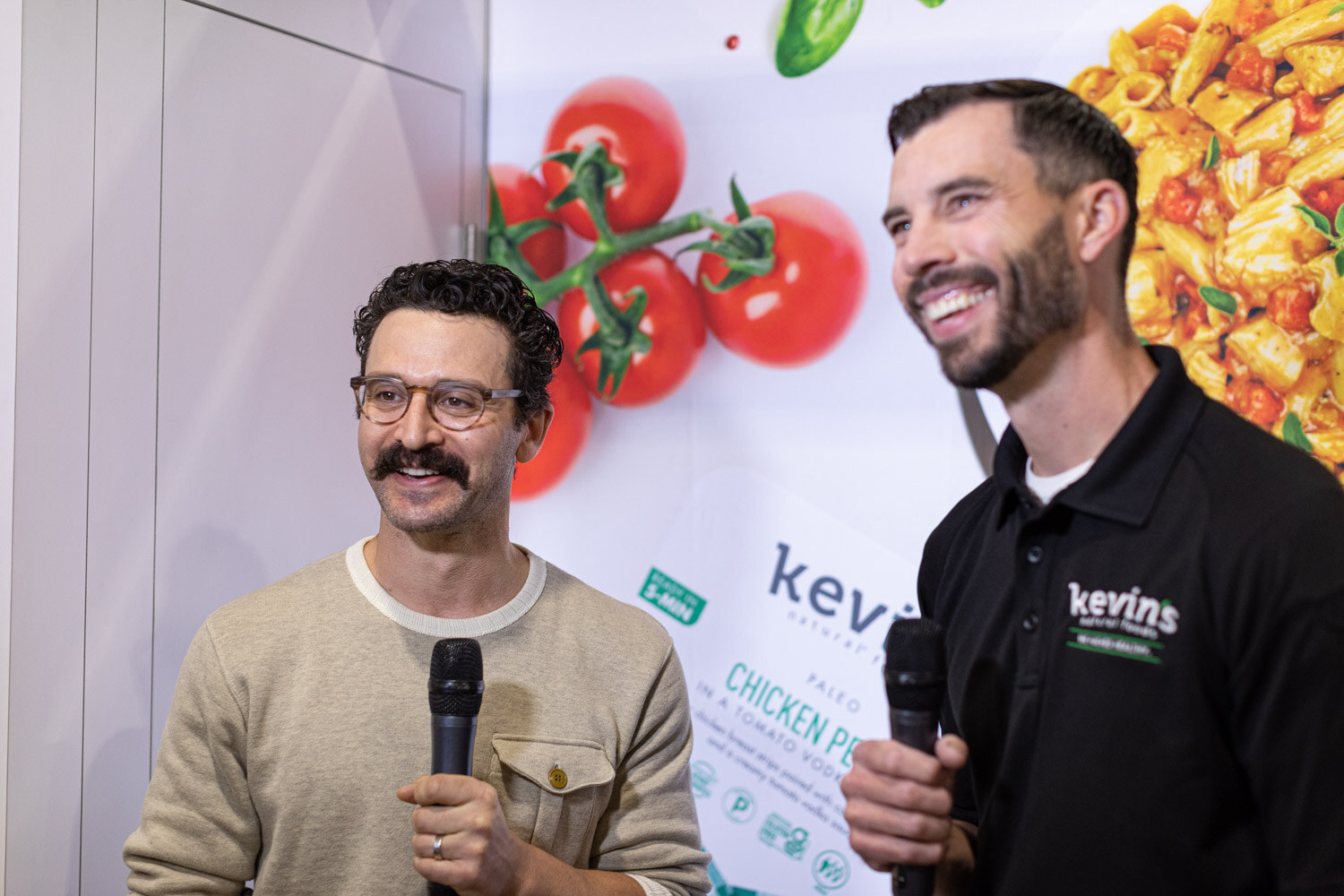 Two men hold microphones in front of a Kevin's Natural Foods banner with tomatoes and pasta; one wears a black Kevin's polo and both are smiling
