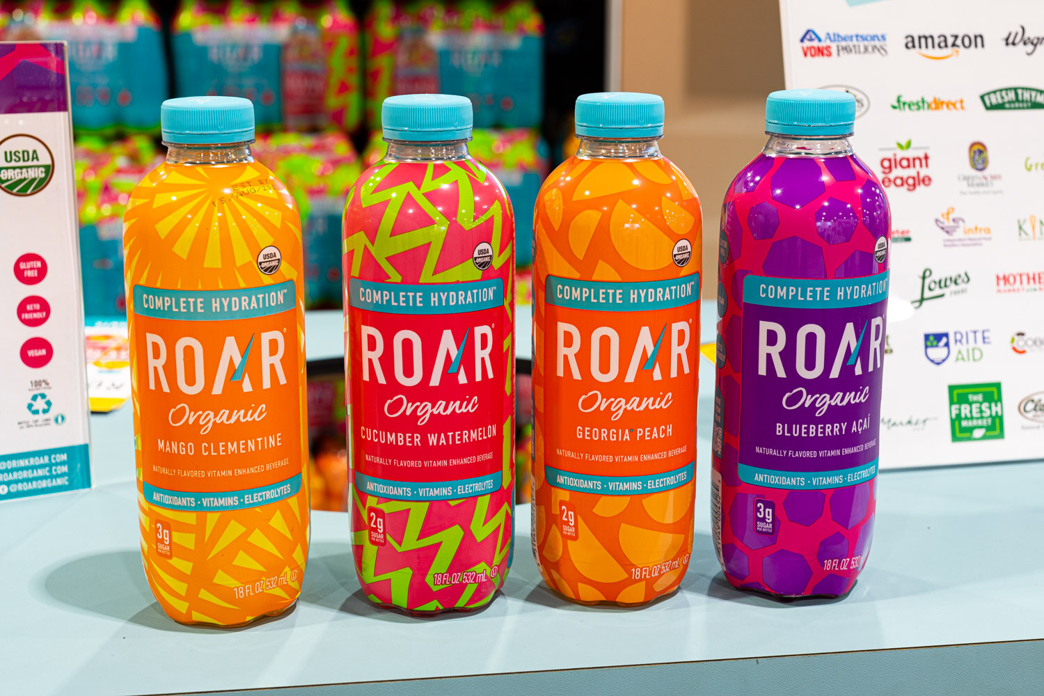 Four colorful ROAR Organic bottles in mango clementine, cucumber watermelon, Georgia peach and blueberry acai stand in front of retailer logos