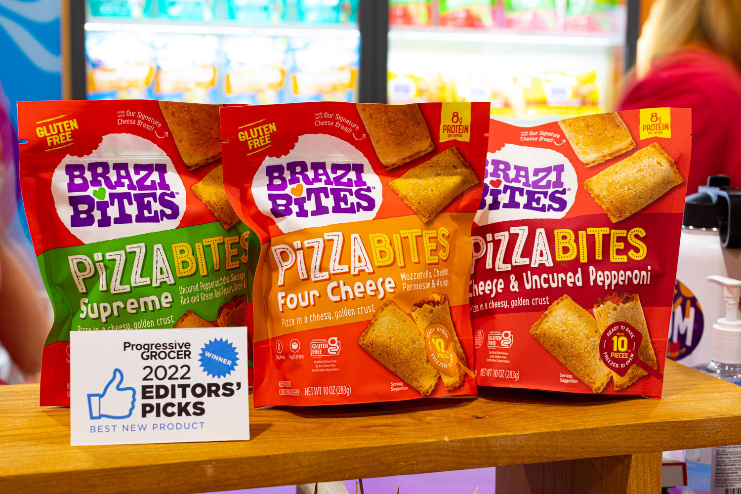 Three Brazi Bites PizzaBites packs stand on a wooden shelf beside a Progressive Grocer 2022 Editors' Picks sign