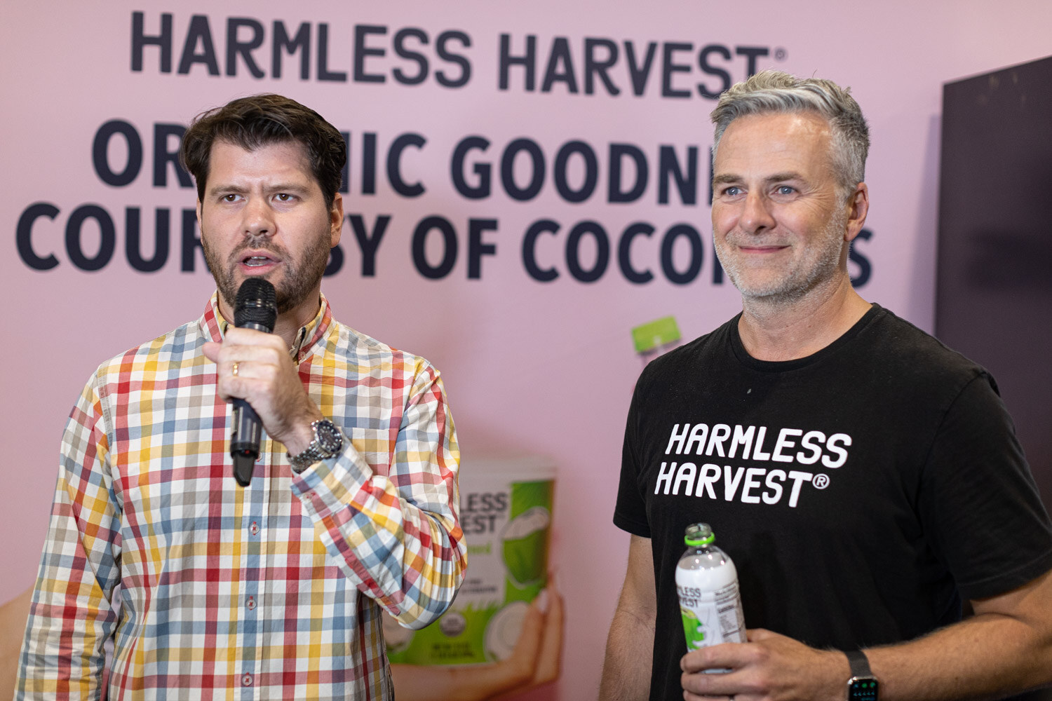 A man in a plaid shirt speaks into a microphone beside a man in a Harmless Harvest t-shirt holding a coconut water bottle in front of a pink Harmless Harvest wall