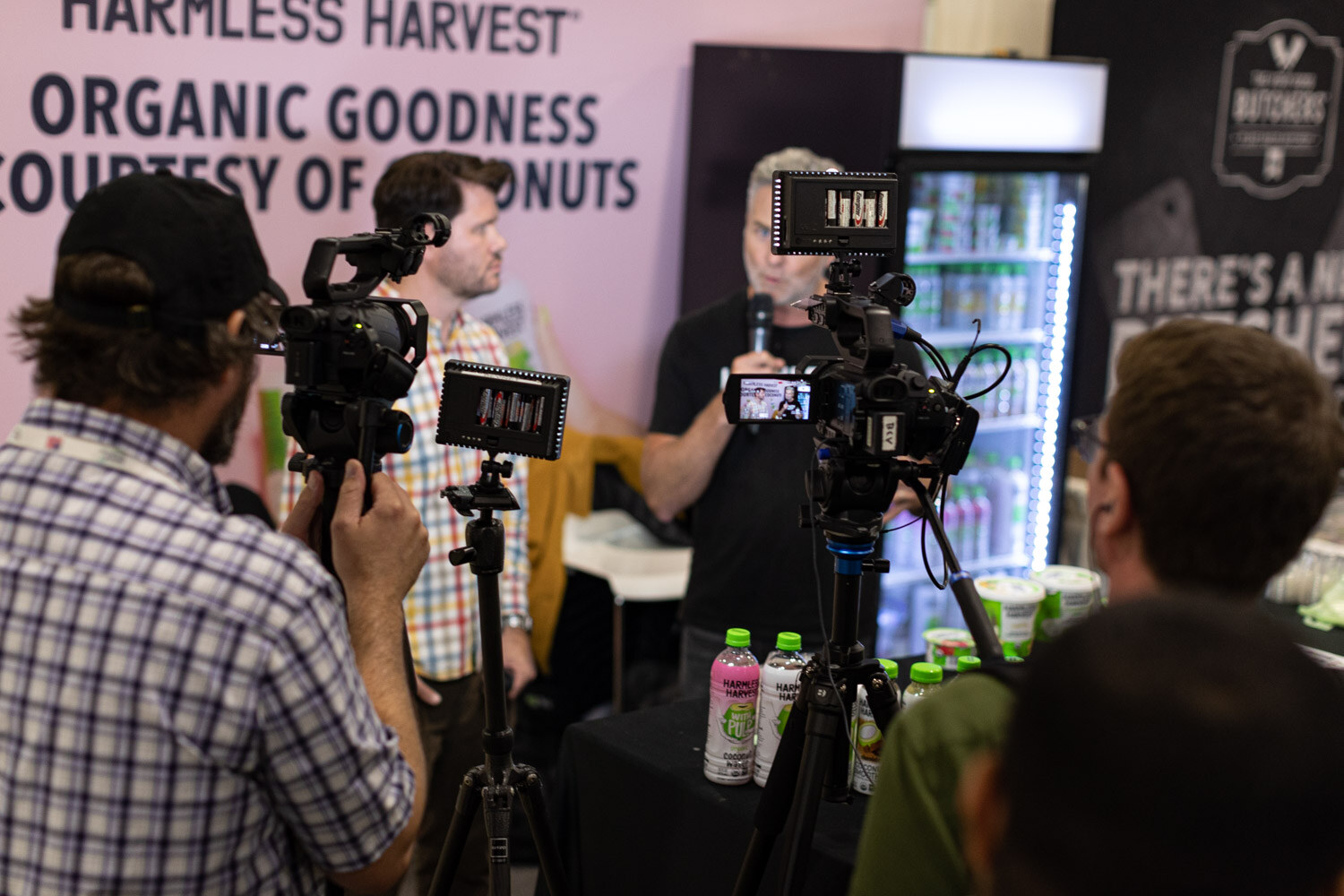 Two camera operators film an interview at the Harmless Harvest booth, with the video screens and a lit cooler in the background