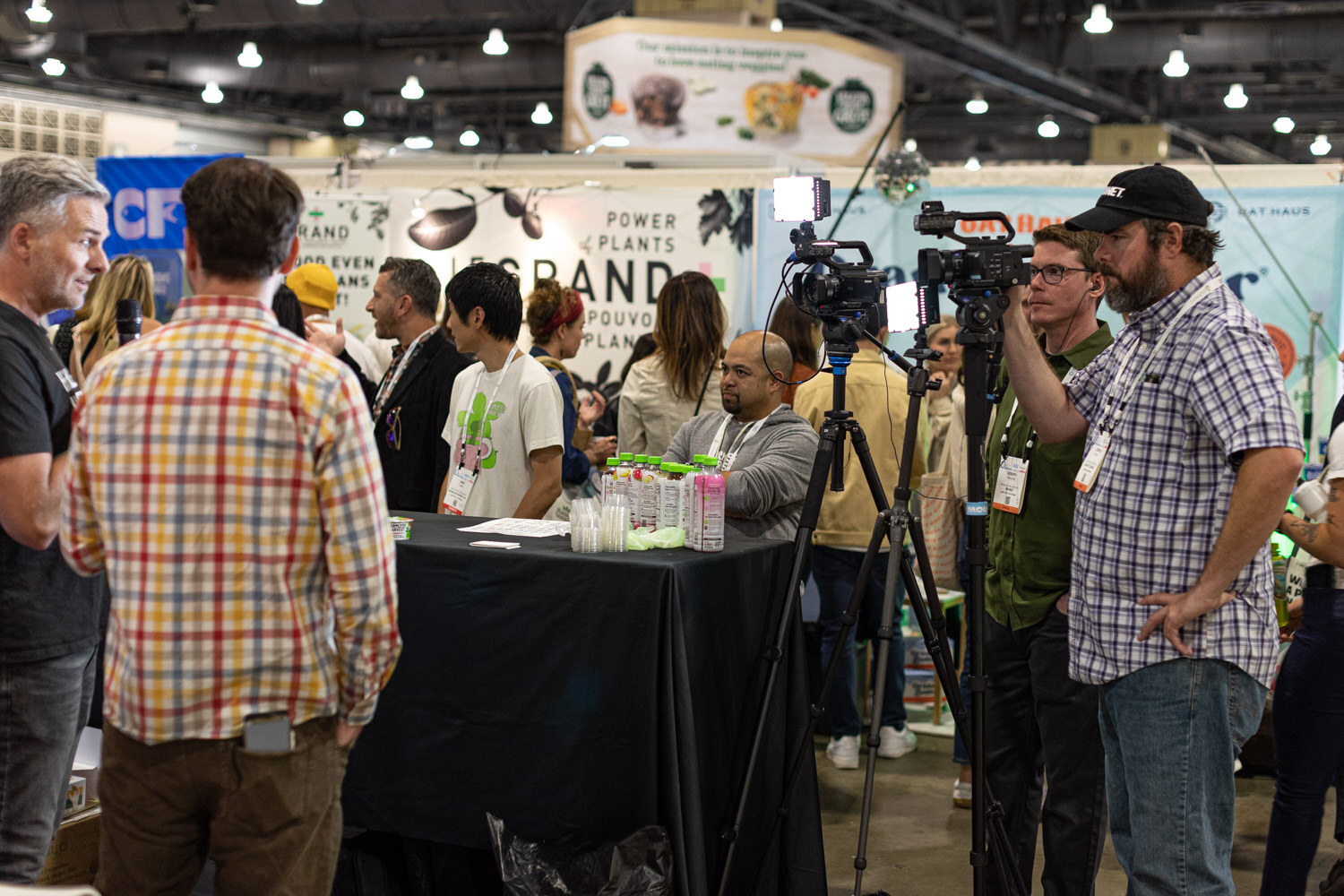 A crowd gathers around a black-draped table while camera operators with tripods and badges record an interview in an exhibit hall aisle