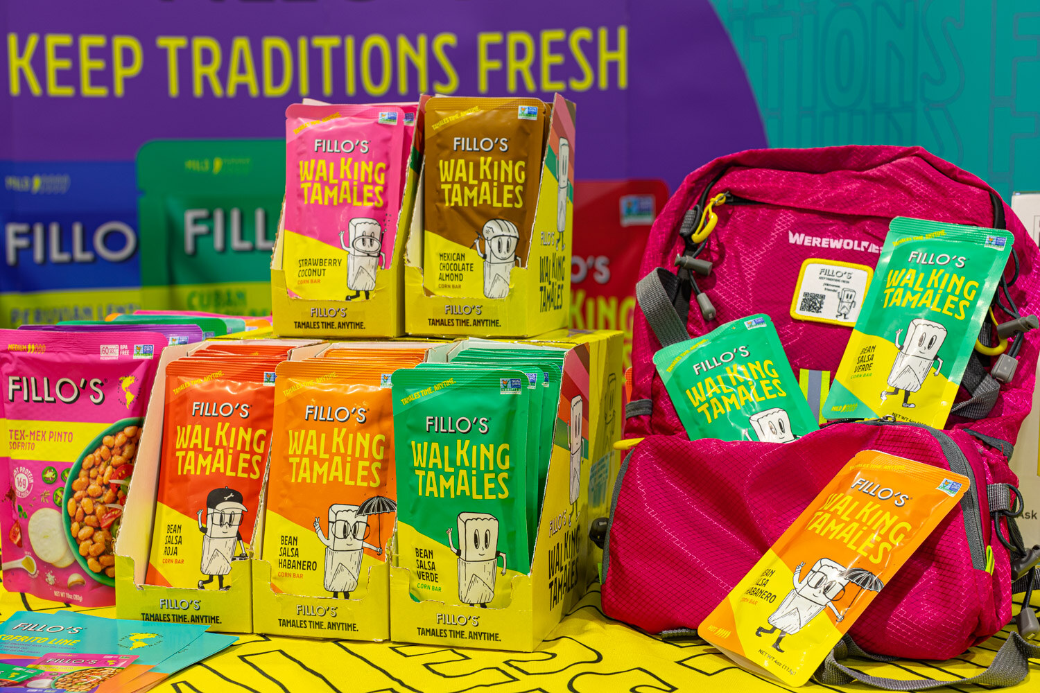 Packages of Fillo's Walking Tamales sit beside a pink backpack on a yellow patterned table under a keep traditions fresh banner