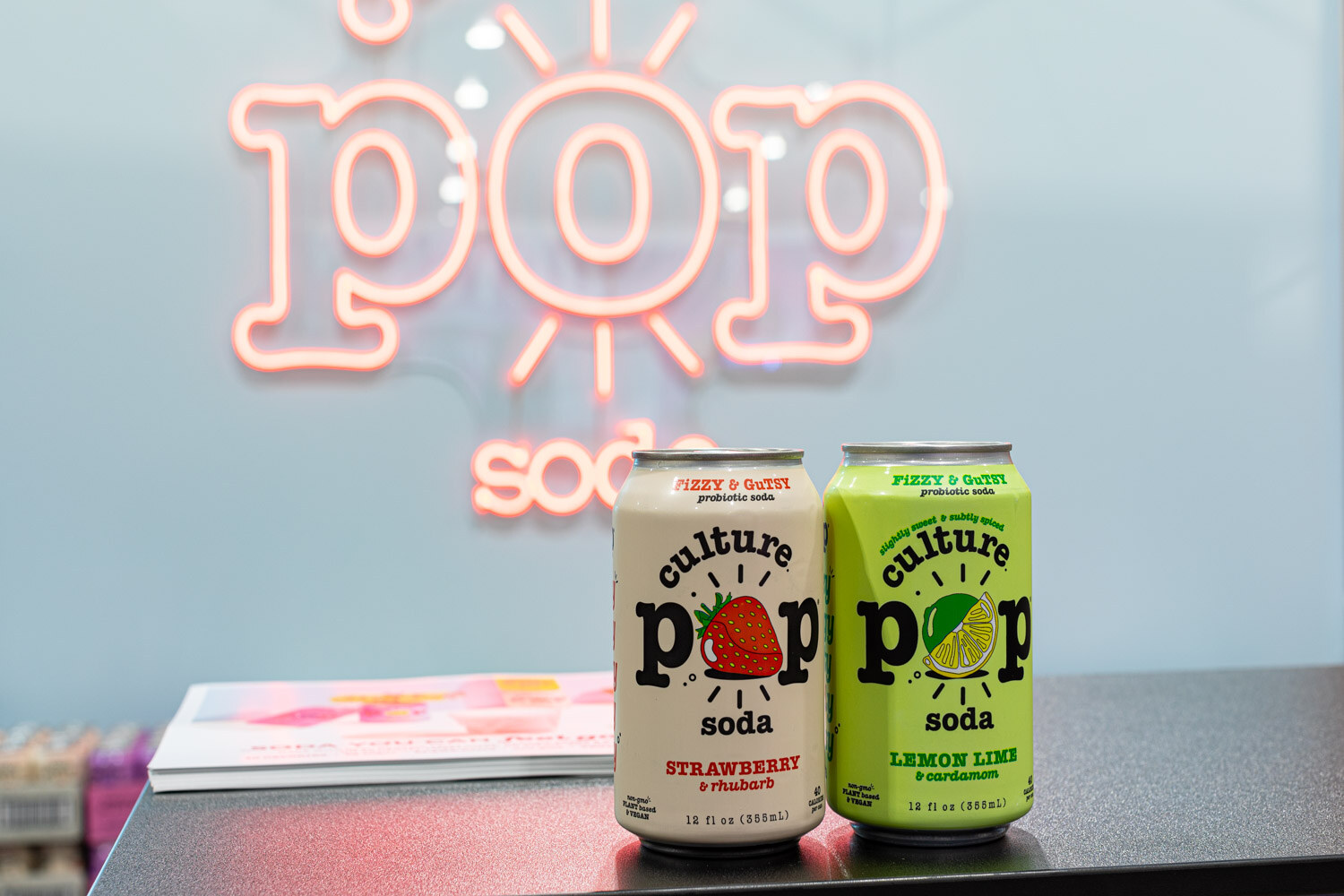 Culture Pop strawberry and lemon lime soda cans stand on a counter in front of a pink neon pop soda sign