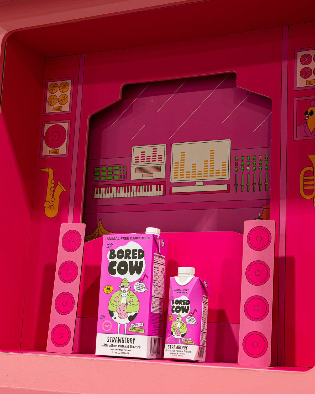 Bored Cow strawberry milk cartons sit inside a bright pink stage-style display with a keyboard and saxophone illustrated on the back