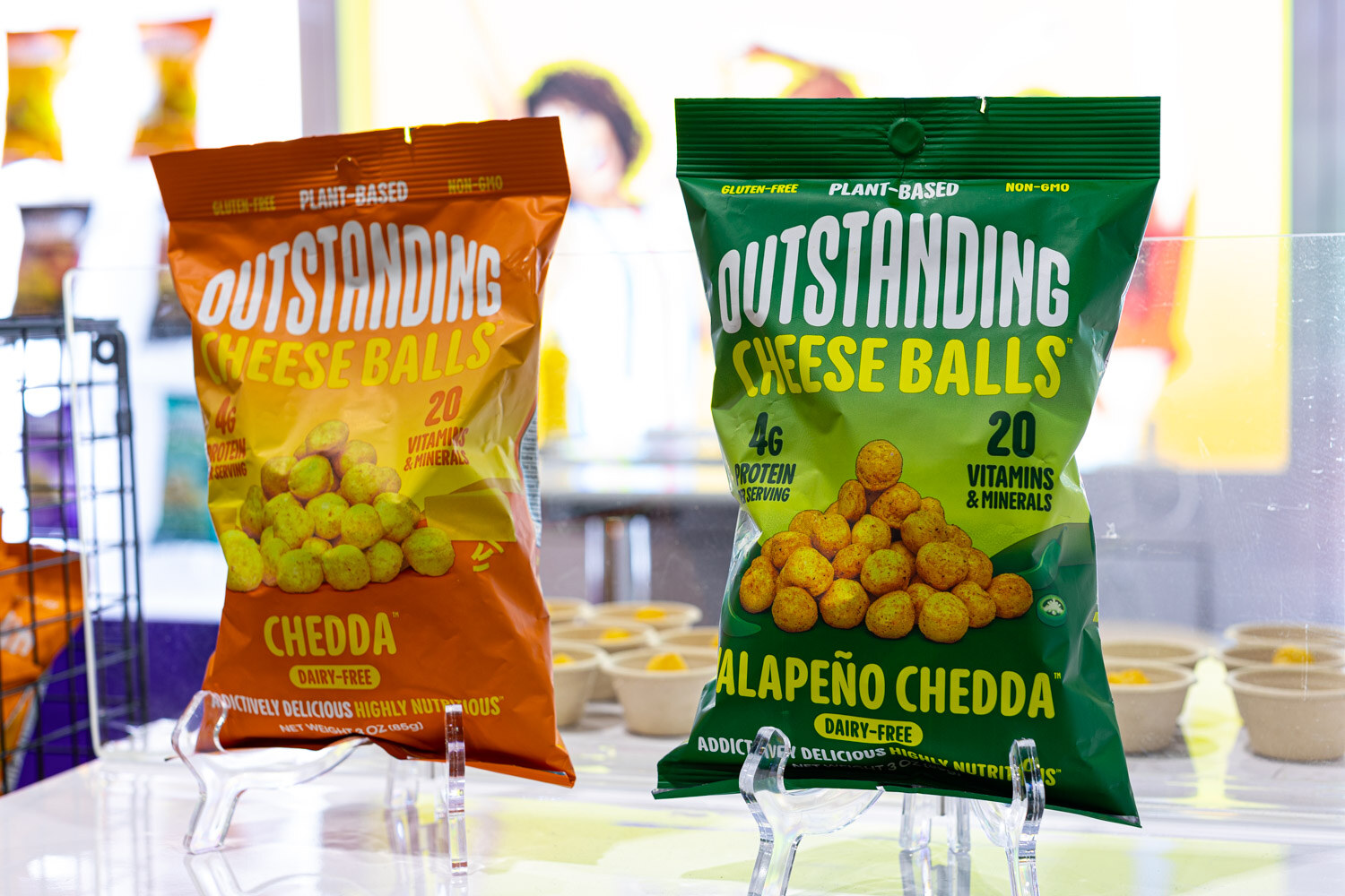 Two Outstanding Cheese Balls bags, Chedda and Jalapeno Chedda, stand on small acrylic stands above sample cups