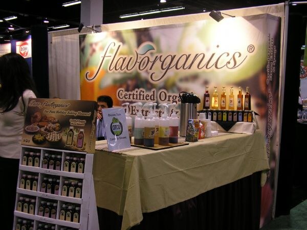 Flavorganics booth with a banner reading certified organic extracts, a table with syrup pump bottles and bottles on a shelf, and a holiday baking display stand in front. A person stands at left.