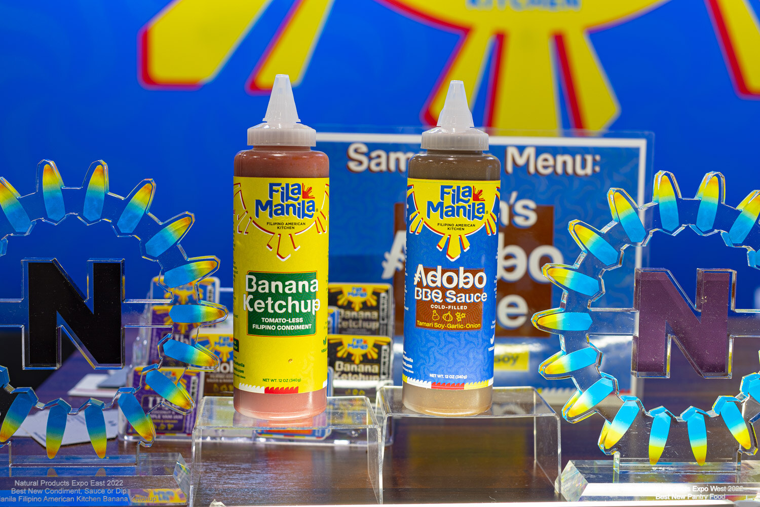 Fila Manila Banana Ketchup and Adobo BBQ Sauce squeeze bottles stand between Fila Manila pouches on clear risers with a Nexty sign behind