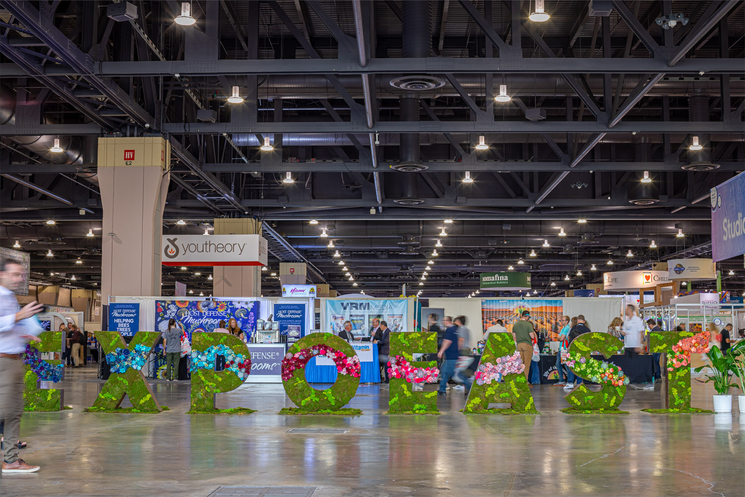 Large green floral letters spelling Expo East stand on the exhibit hall floor with attendees and booths behind them