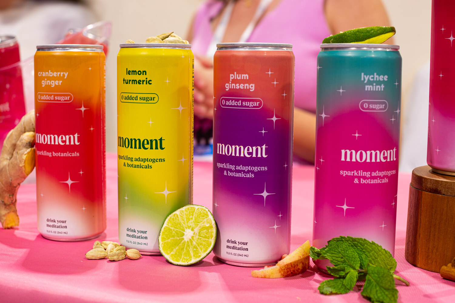 Four Moment cans in cranberry ginger, lemon turmeric, plum ginseng and lychee mint flavors with a lime and mint on a pink table