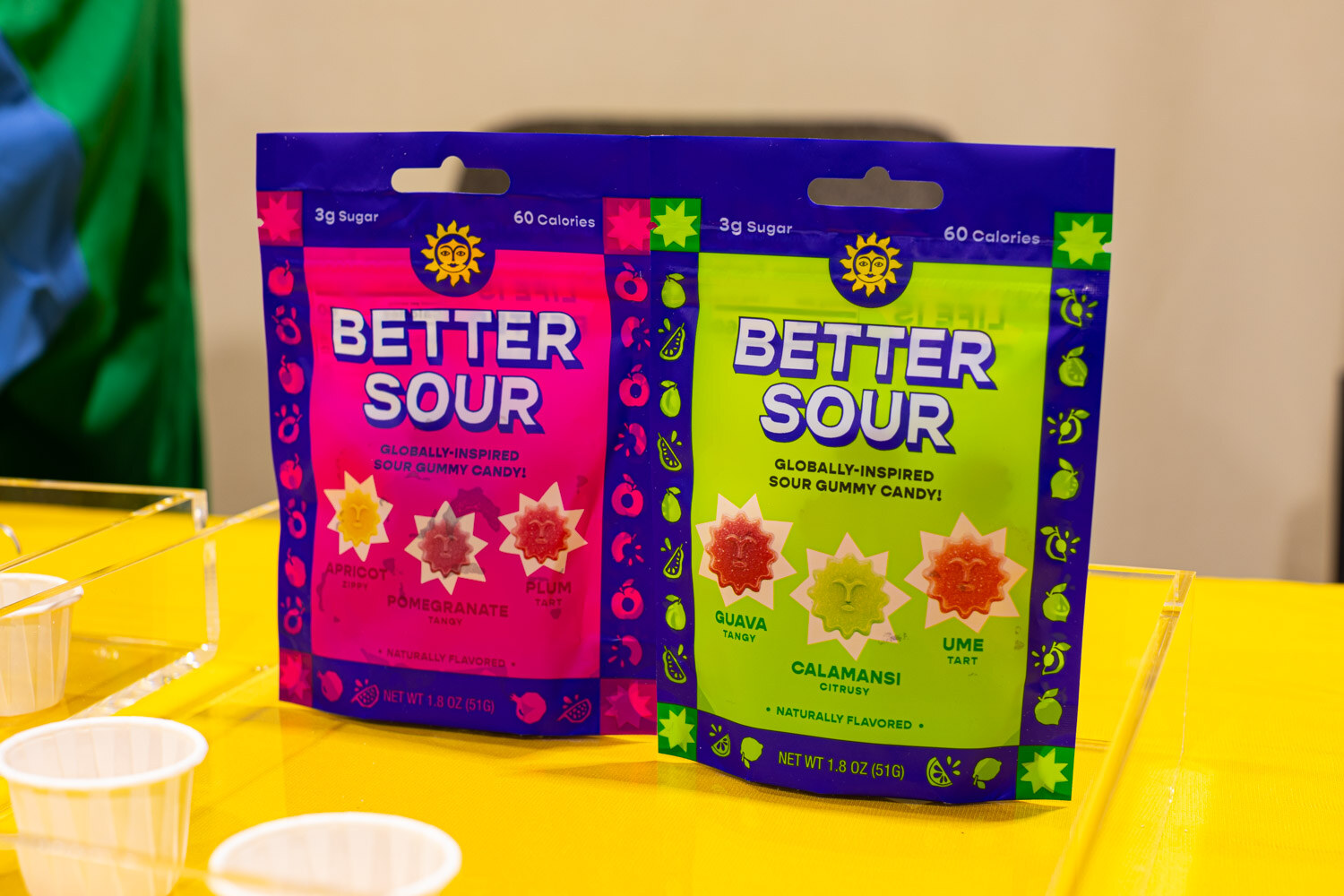 Two bright pink and green Better Sour sour gummy pouches stand on a yellow table with sample cups beside them