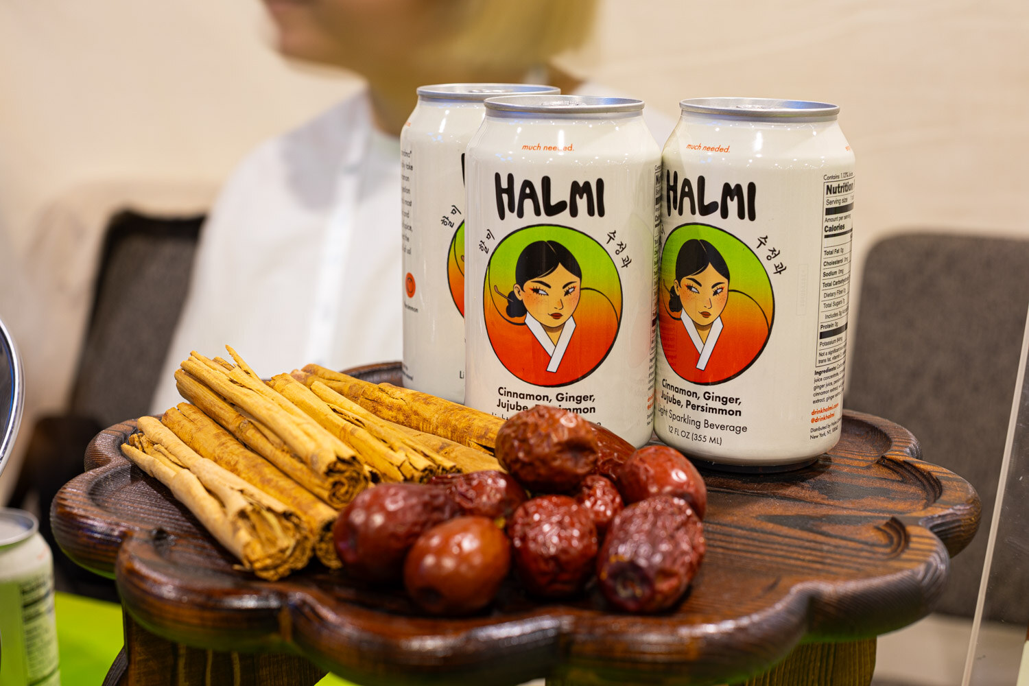 Two Halmi cans with an illustrated woman on the label sit on a wooden tray with cinnamon sticks and dried dates
