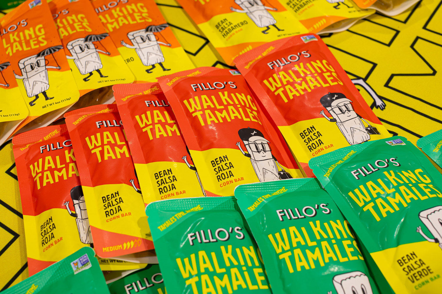 Piles of Fillo's Walking Tamales in bean salsa roja and bean salsa verde packaging on a yellow patterned table