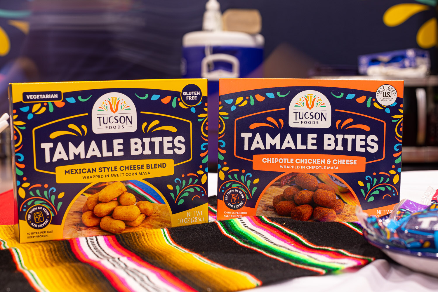 Two Tucson Foods Tamale Bites boxes, Mexican Style Cheese Blend and Chipotle Chicken and Cheese, sit on a striped serape