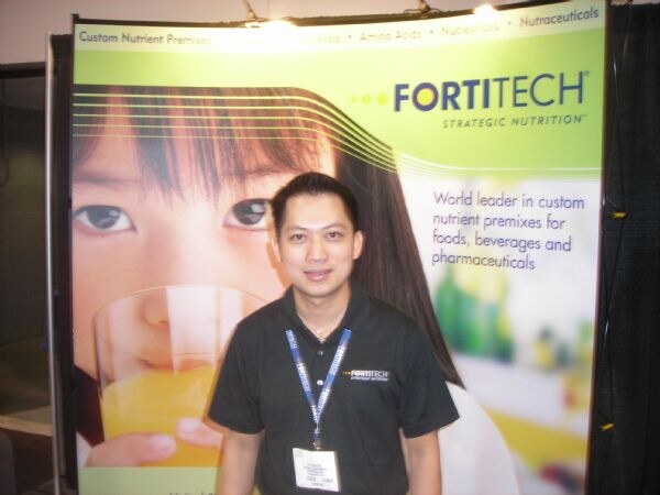 A smiling man in a black Fortitech polo and lanyard stands in front of a Fortitech banner showing a child drinking orange juice and text about custom nutrient premixes.