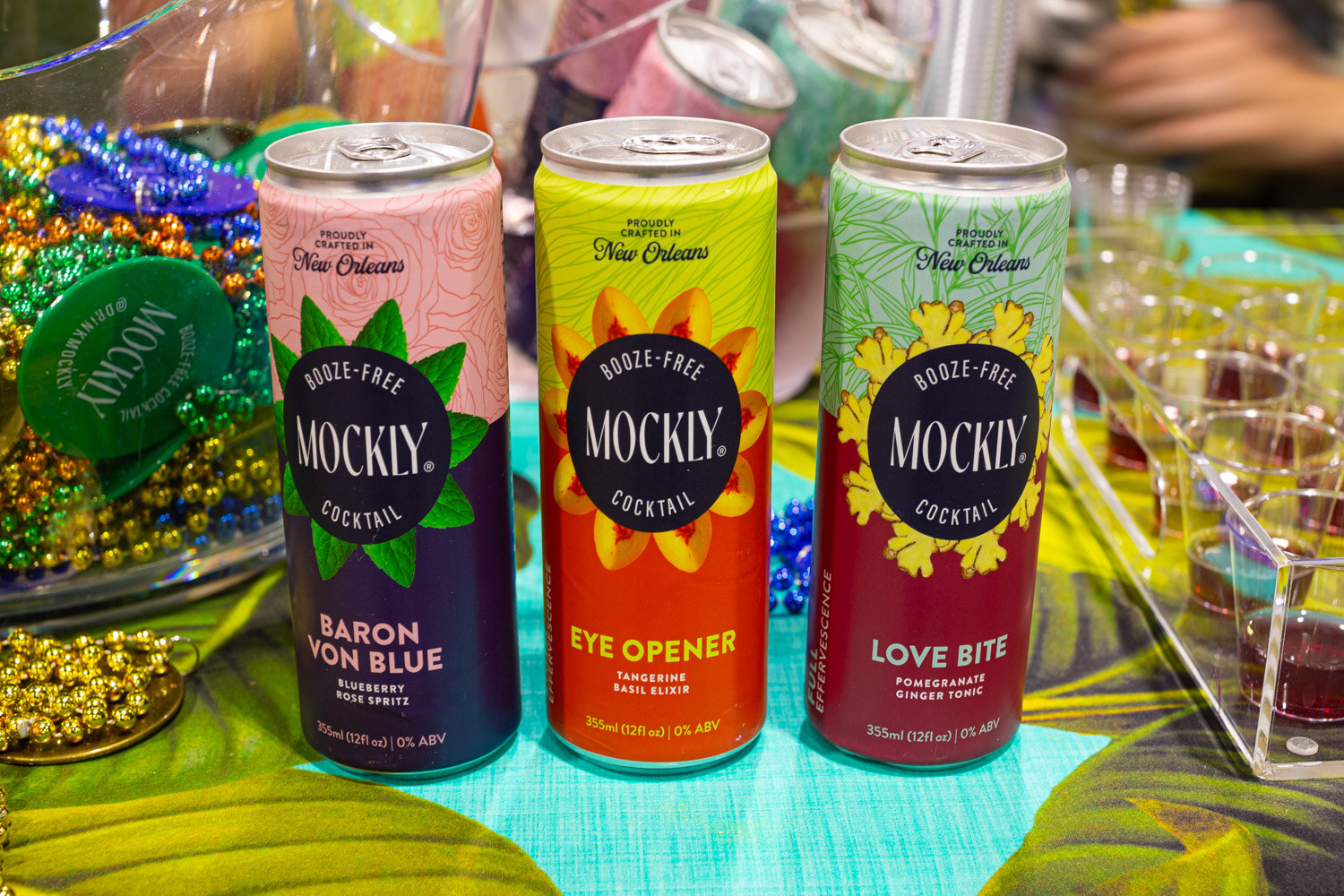 Three Mockly cans, Baron Von Blue, Eye Opener and Love Bite, sit among beads on a bright New Orleans themed table
