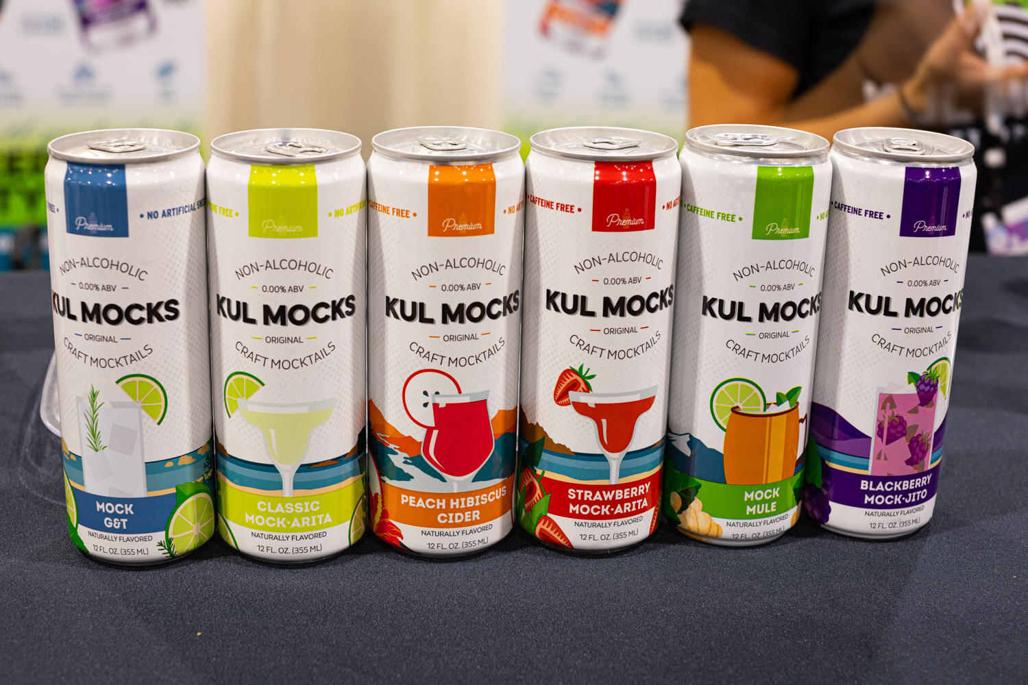 Six Kul Mocks non-alcoholic craft mocktail cans in lime, red and purple labels sit in a row on a navy tablecloth