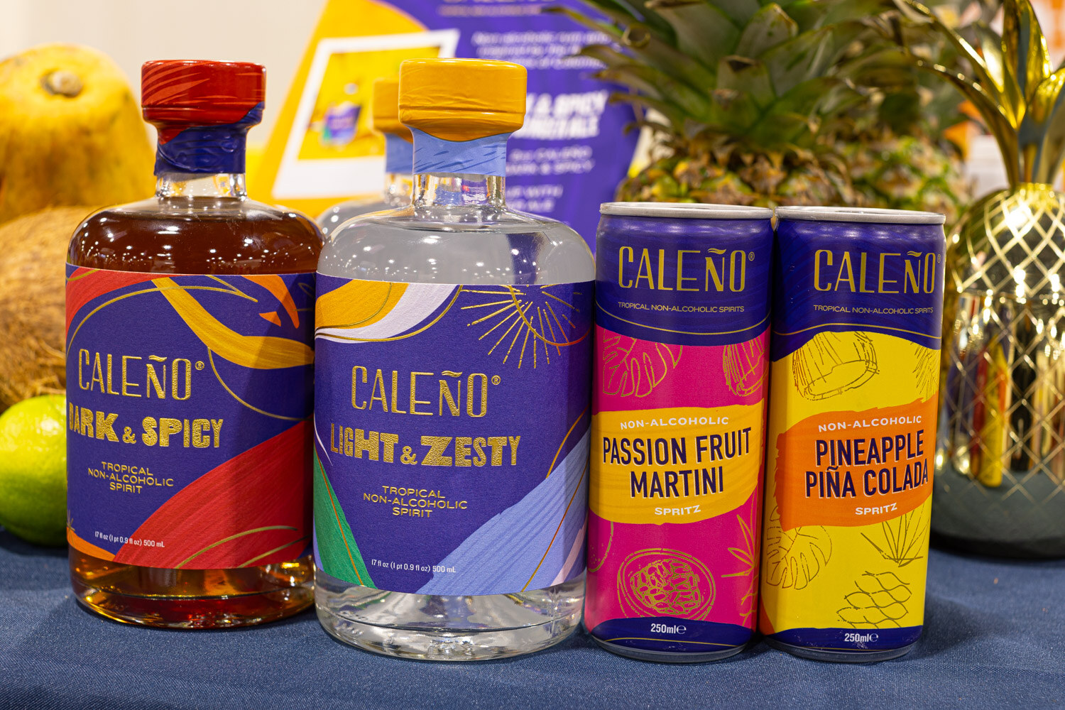 Bottles of Caleno Dark and Spicy and Light and Zesty stand beside Caleno Passion Fruit Martini and Pineapple Pina Colada cans on a pineapple-decorated table