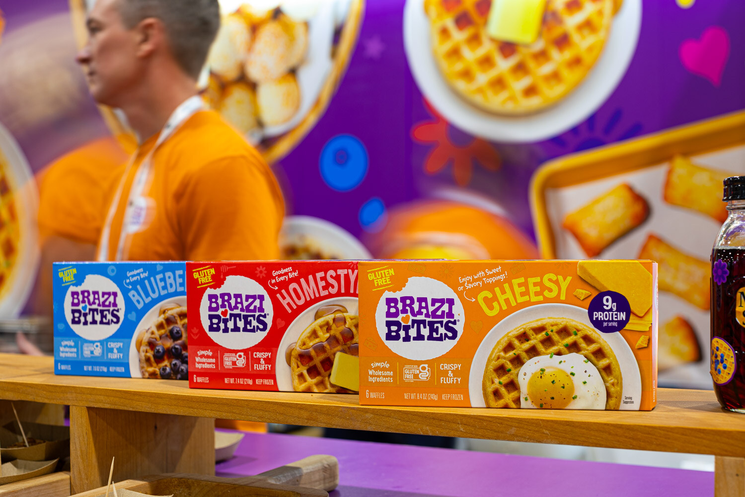 Boxes of Brazi Bites in blueberry waffle, homestyle and cheesy flavors sit on a wooden shelf in front of a purple display
